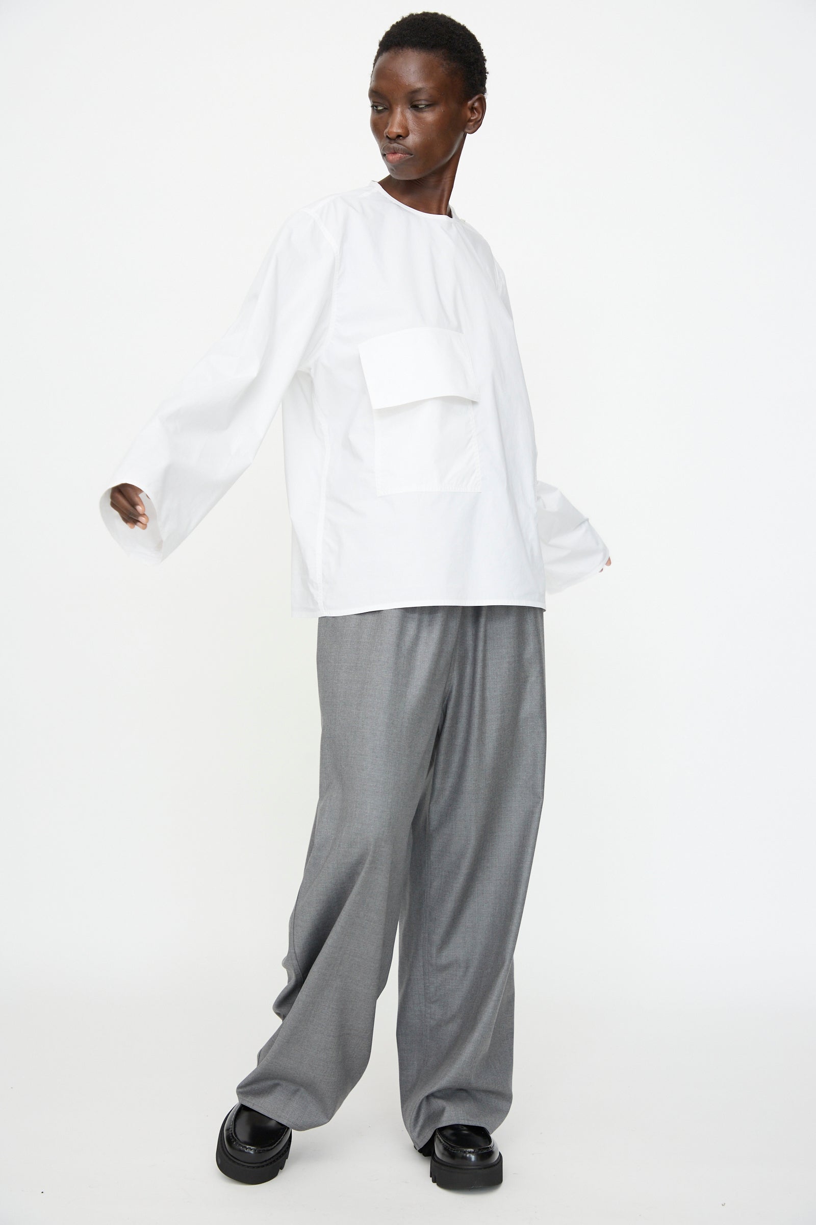 Model wears the Sofie D'Hoore Cotton Poplin Balthus Shirt in White, featuring an oversized fit and large front pockets, paired with wide gray trousers and black shoes, standing against a plain white background.