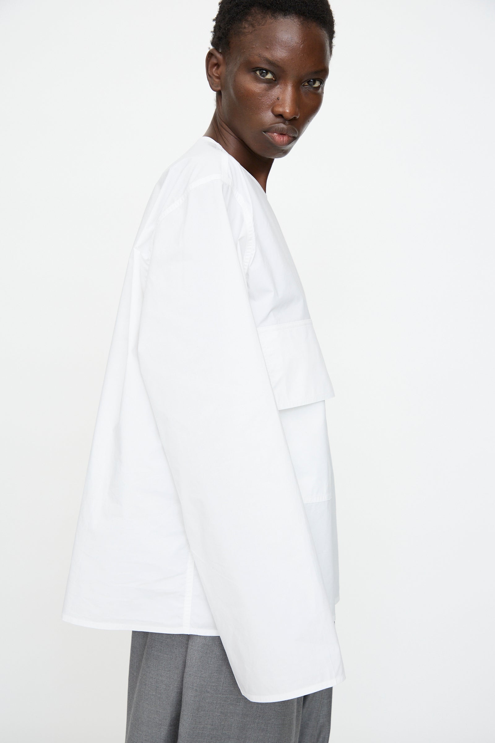 A person with short hair wears an oversized fit Sofie D'Hoore Cotton Poplin Balthus Shirt in White and gray pants, standing sideways against a plain white background.