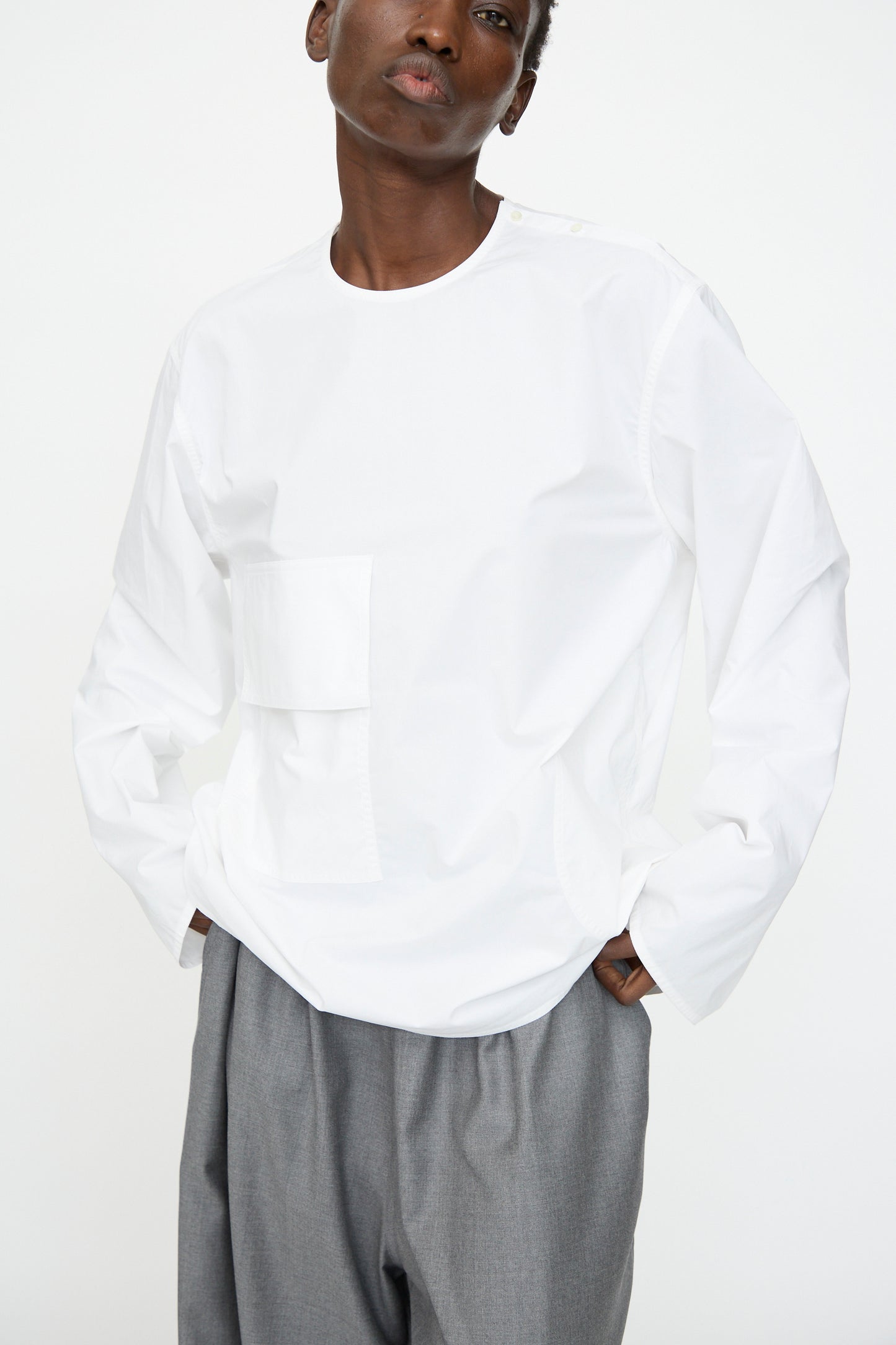 A person wears a Sofie D'Hoore Cotton Poplin Balthus Shirt in white, featuring an oversized fit, front pocket, and long sleeves, paired with gray trousers, standing against a plain white background.
