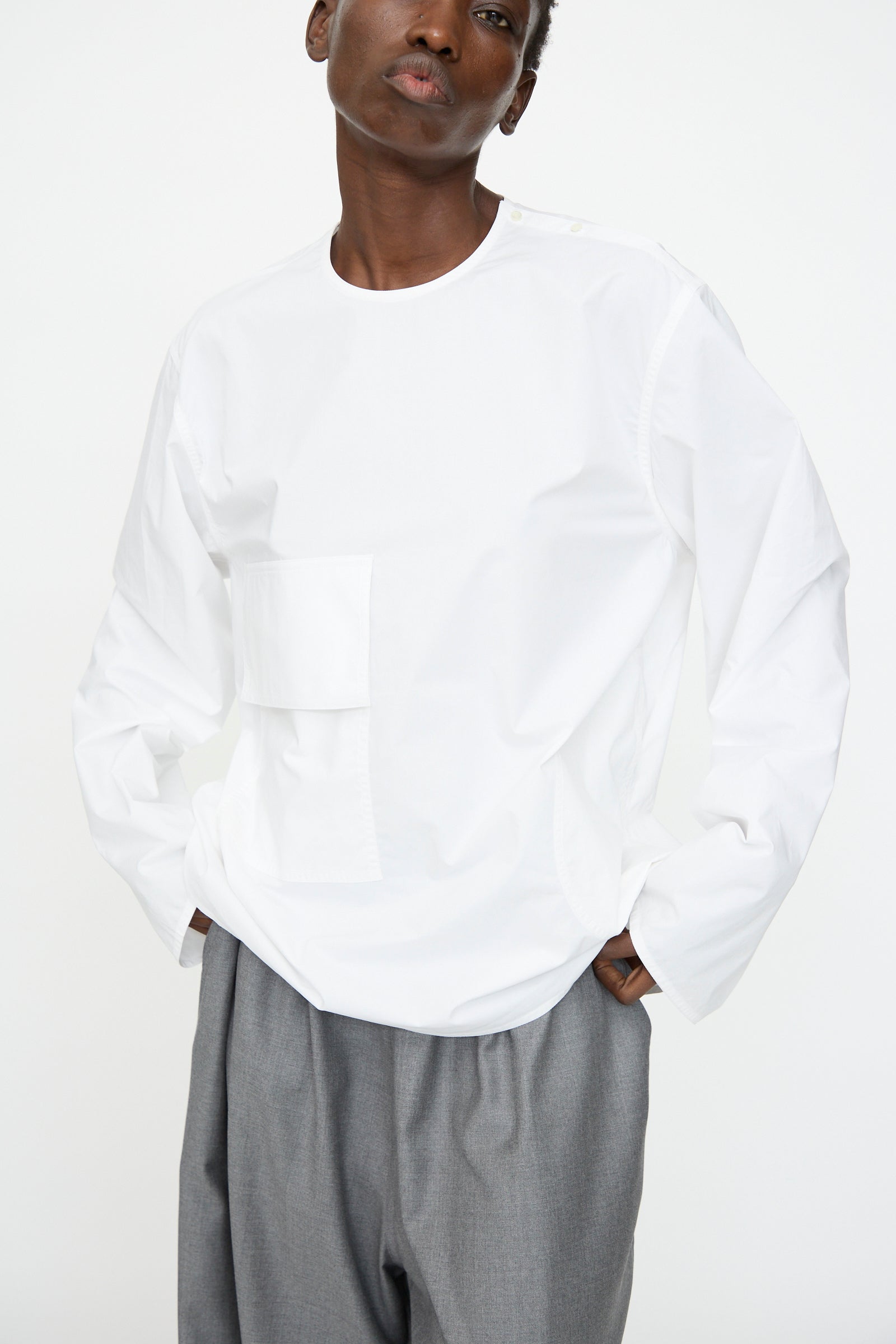 A person wears a Sofie D'Hoore Cotton Poplin Balthus Shirt in white, featuring an oversized fit, front pocket, and long sleeves, paired with gray trousers, standing against a plain white background.