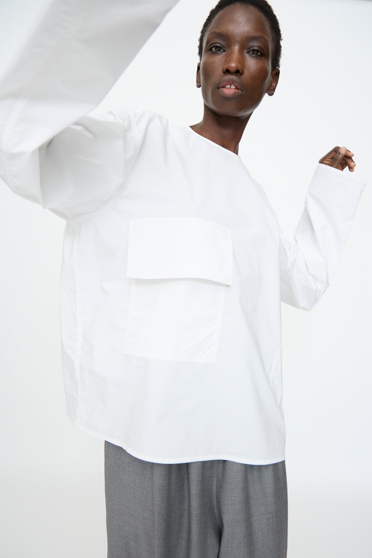 A person wears the Sofie D'Hoore Cotton Poplin Balthus Shirt in White—an oversized cotton poplin shirt with a large front pocket—paired with gray pants, posing against a plain white background.