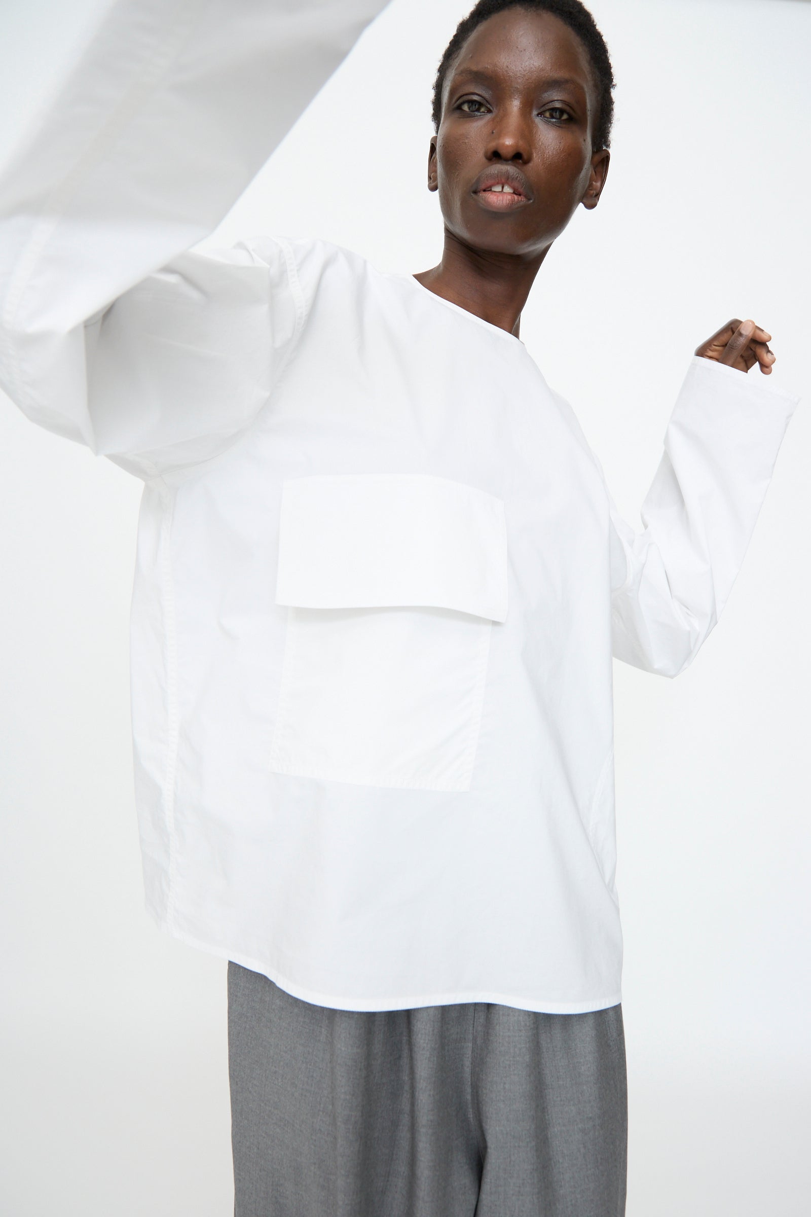 A person wears the Sofie D'Hoore Cotton Poplin Balthus Shirt in White—an oversized cotton poplin shirt with a large front pocket—paired with gray pants, posing against a plain white background.