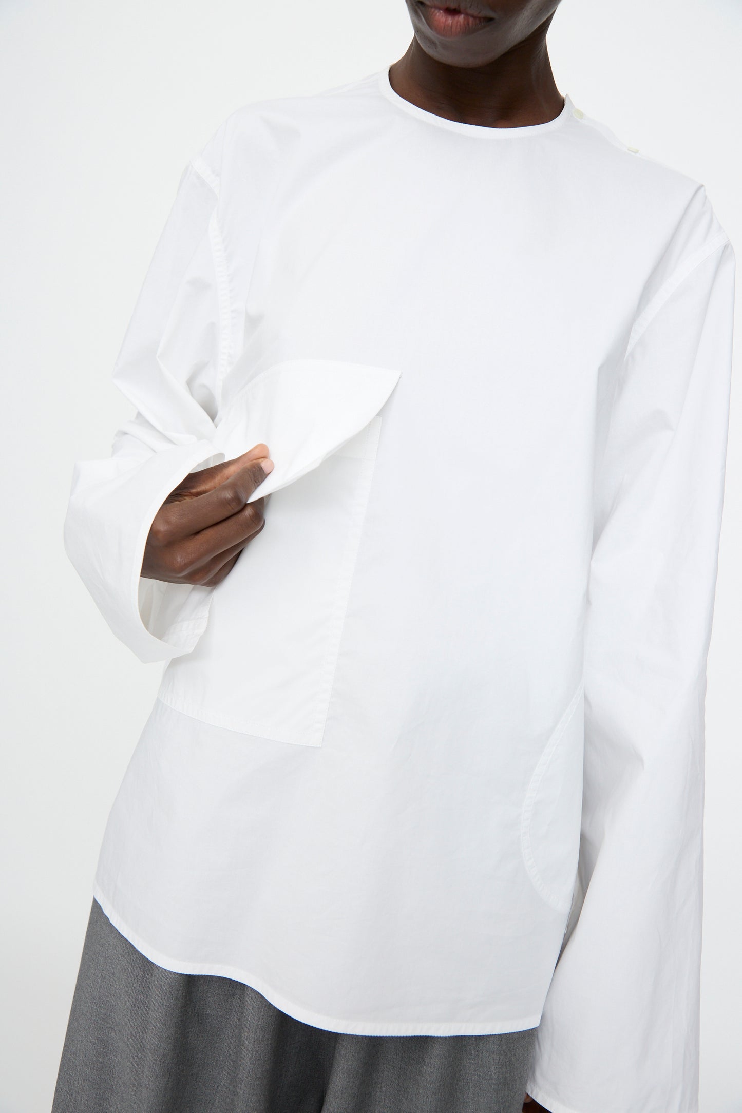 A person wearing an oversized Sofie D'Hoore Cotton Poplin Balthus Shirt in White with wide sleeves and gray pants, holding the shirt's flap pocket.