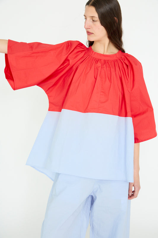 A person in an oversize fit outfit, wearing the Sofie D'Hoore Cotton Poplin Barcelona Trapeze Top in Poppy and Sky, extends an arm to the side.