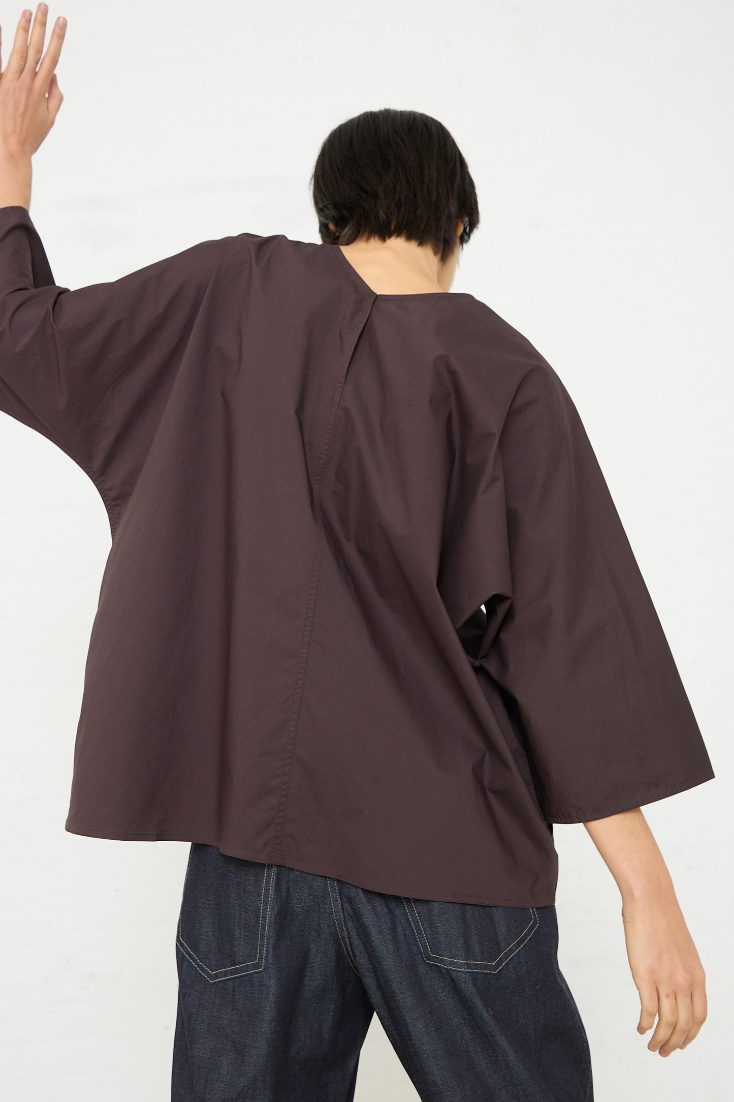 A woman in a brown shirt with an oversized fit, the Sofie D'Hoore Cotton Poplin Bendol Top with Scarf in Ebony, is standing with her arms outstretched. Back view.