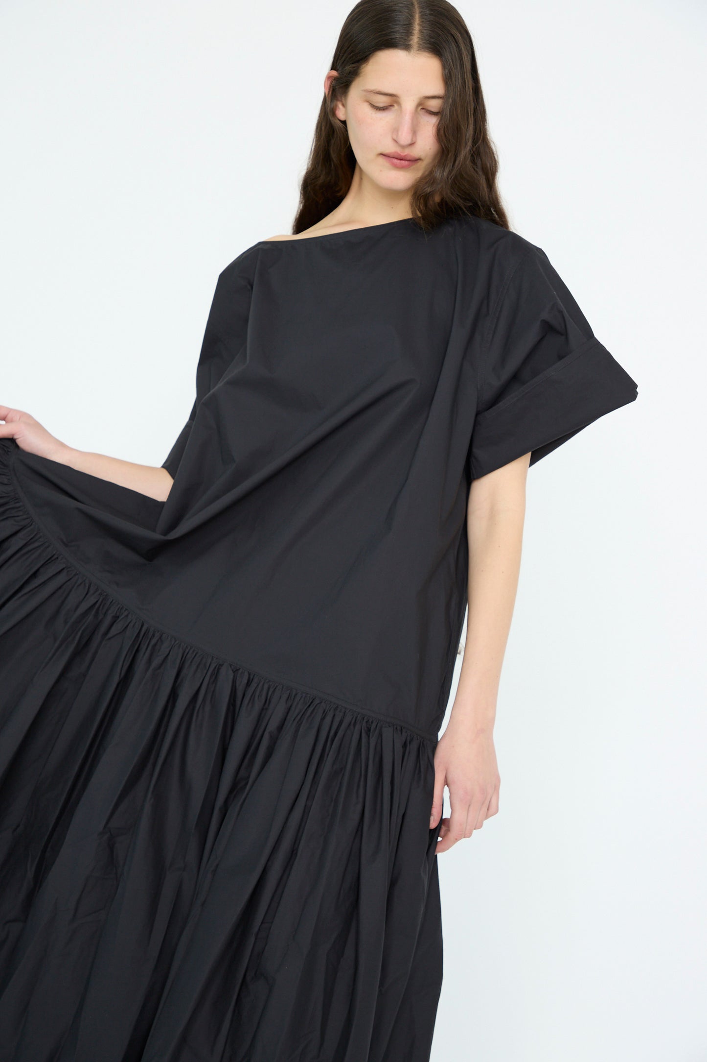 A person holds the hem of a loose-fitting Sofie D'Hoore Cotton Poplin Della Dress in Black, showcasing its short sleeves, oversized design with a gathered skirt and dropped waist, against a plain white background.