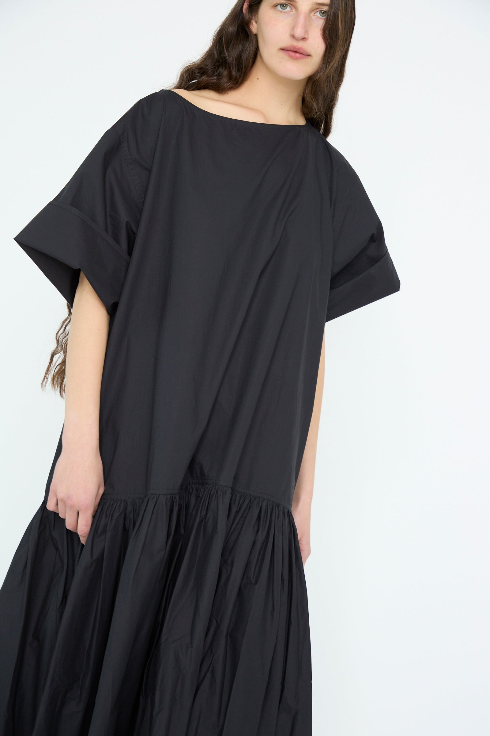 A person is wearing the Sofie D'Hoore Cotton Poplin Della Dress in black, featuring an oversized fit, short sleeves, and a gathered skirt, standing against a plain background.