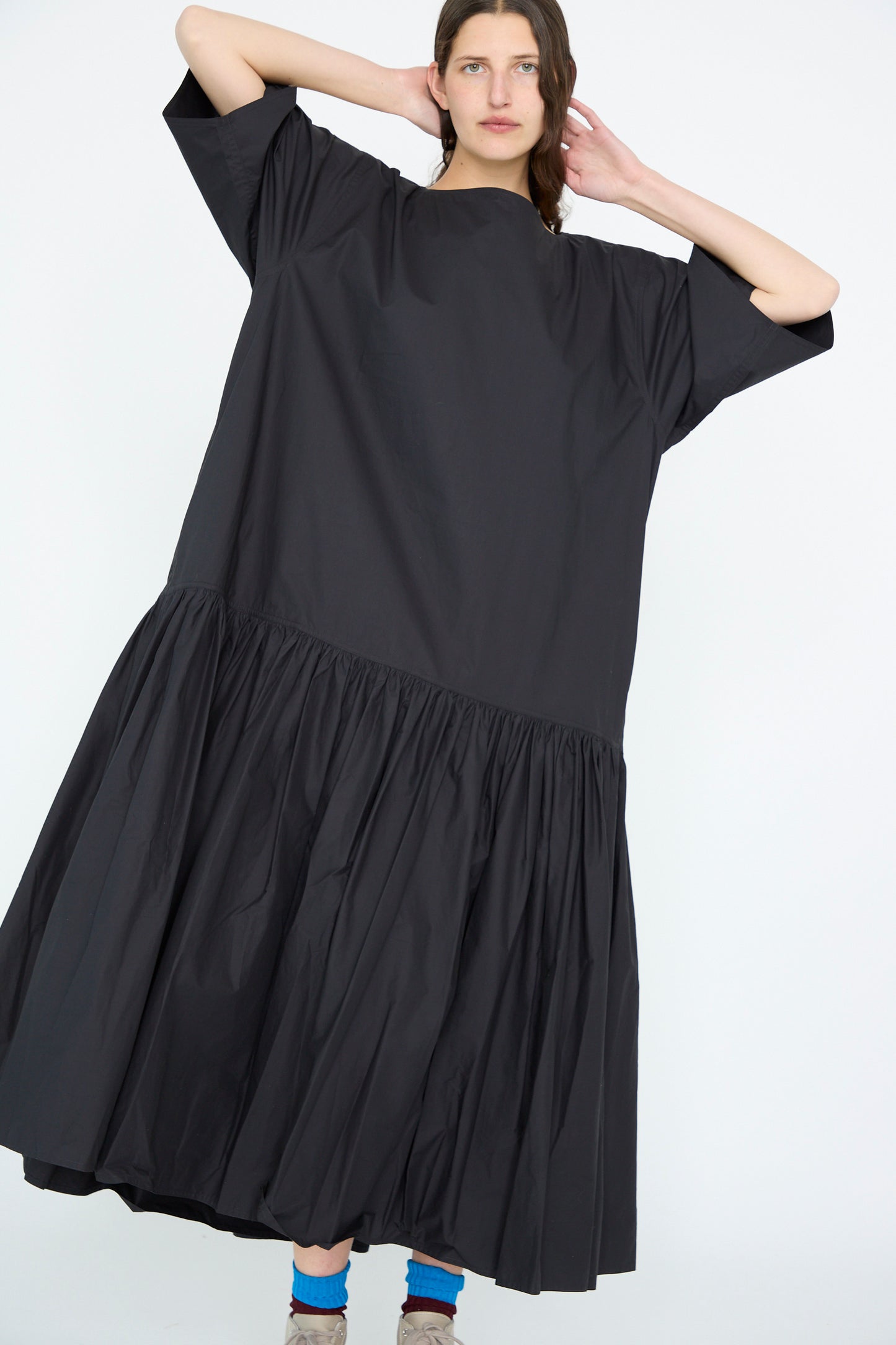 A person poses against a light background wearing the Sofie D'Hoore Cotton Poplin Della Dress in Black, an oversized design with a gathered skirt and dropped waist. One arm is raised, and they wear colorful socks paired with ankle-high shoes.