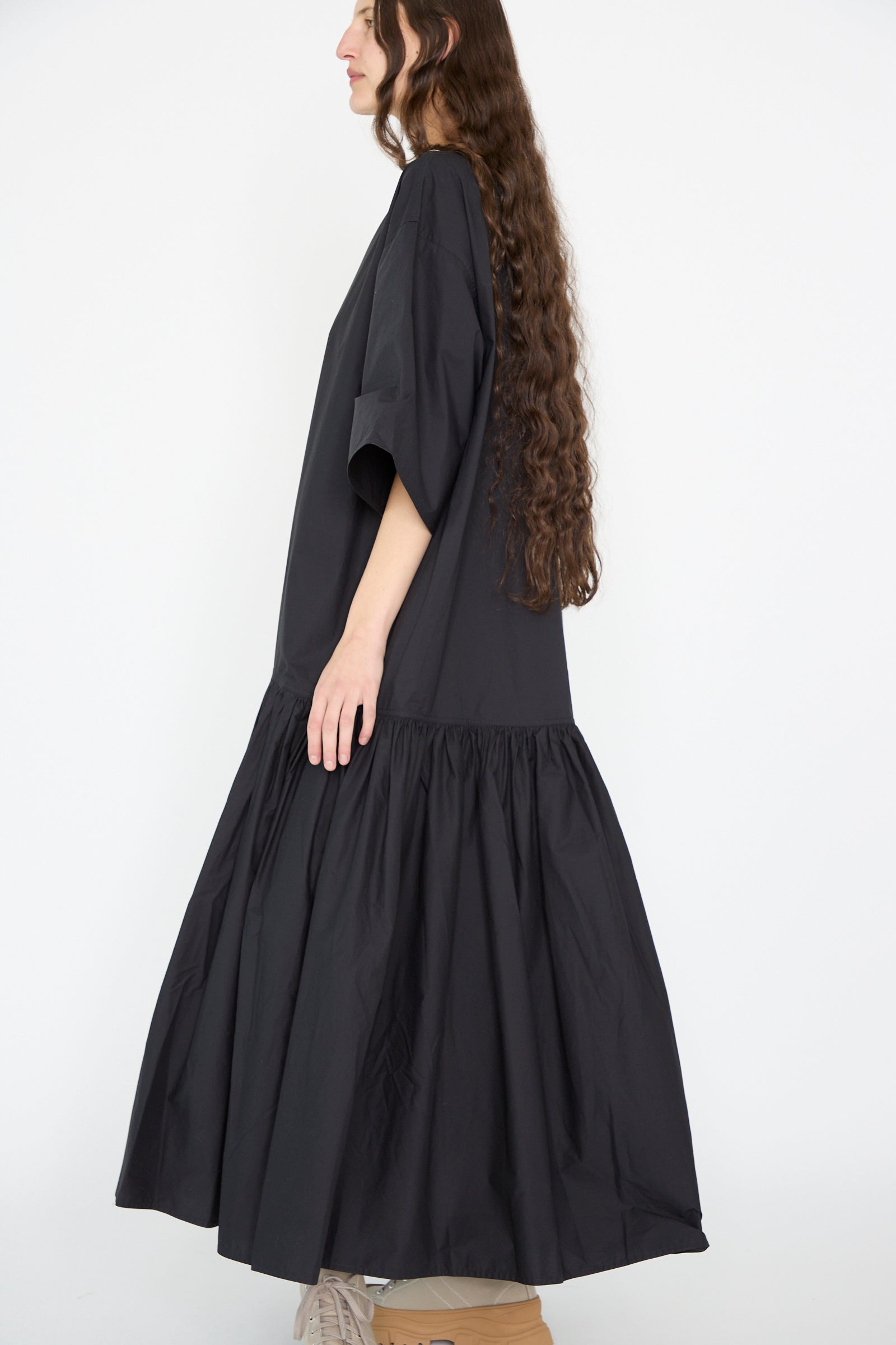 A woman with long, curly hair stands in profile wearing the oversized, ankle-length Cotton Poplin Della Dress in Black by Sofie D'Hoore, featuring a gathered hem.