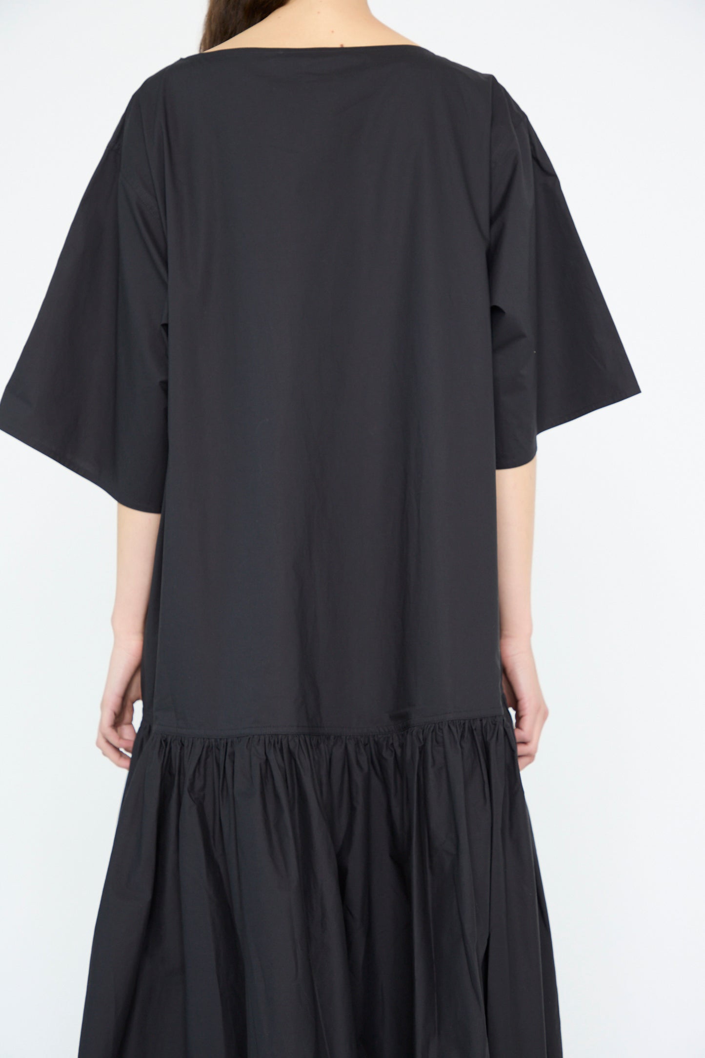 Viewed from the back against a plain backdrop, an individual wears the Cotton Poplin Della Dress in Black by Sofie D'Hoore. This oversized, loose-fitting dress features short sleeves and a gathered skirt.