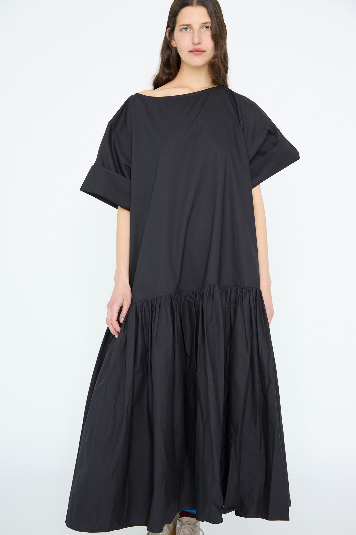 A person stands against a plain backdrop, wearing the oversized Cotton Poplin Della Dress in black by Sofie D'Hoore. The dress features short sleeves and a dropped waist.