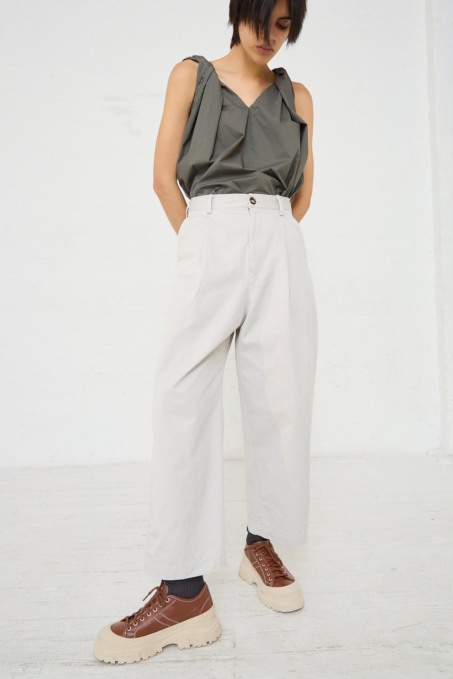 A woman is standing in a white room wearing Sofie D'Hoore's light stone grey Cotton Twill Proof Pant in Mastic with pleat detail.