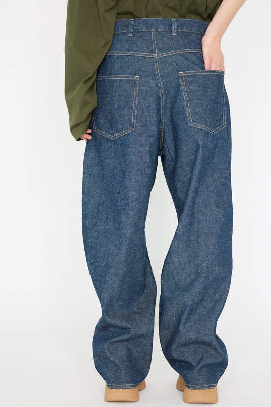 A person stands backward against a white backdrop wearing Sofie D'Hoore's oversized "Denim Peggy Jean in Blue" with a green long-sleeve top, their hand resting casually in the rear pocket of the cotton and hemp blend jeans.