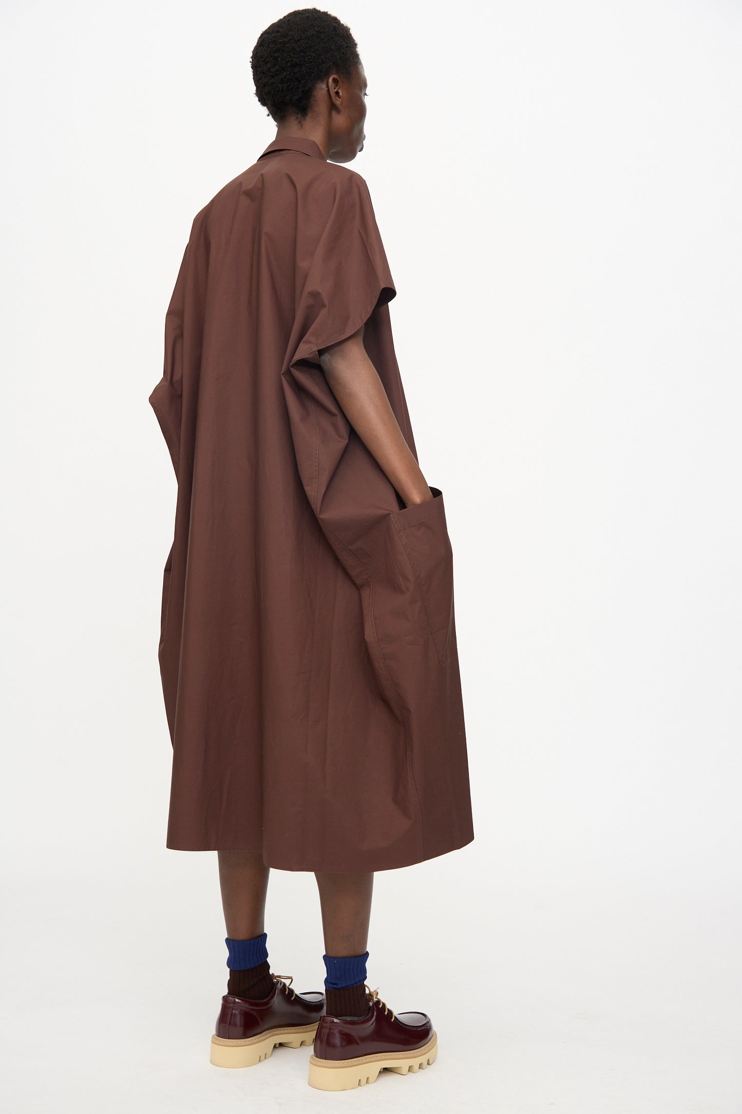 A person stands sideways against a white background, wearing the oversized fit Sofie D'Hoore Duchy Dress in Cacao with short sleeves, styled with blue socks and brown platform shoes.