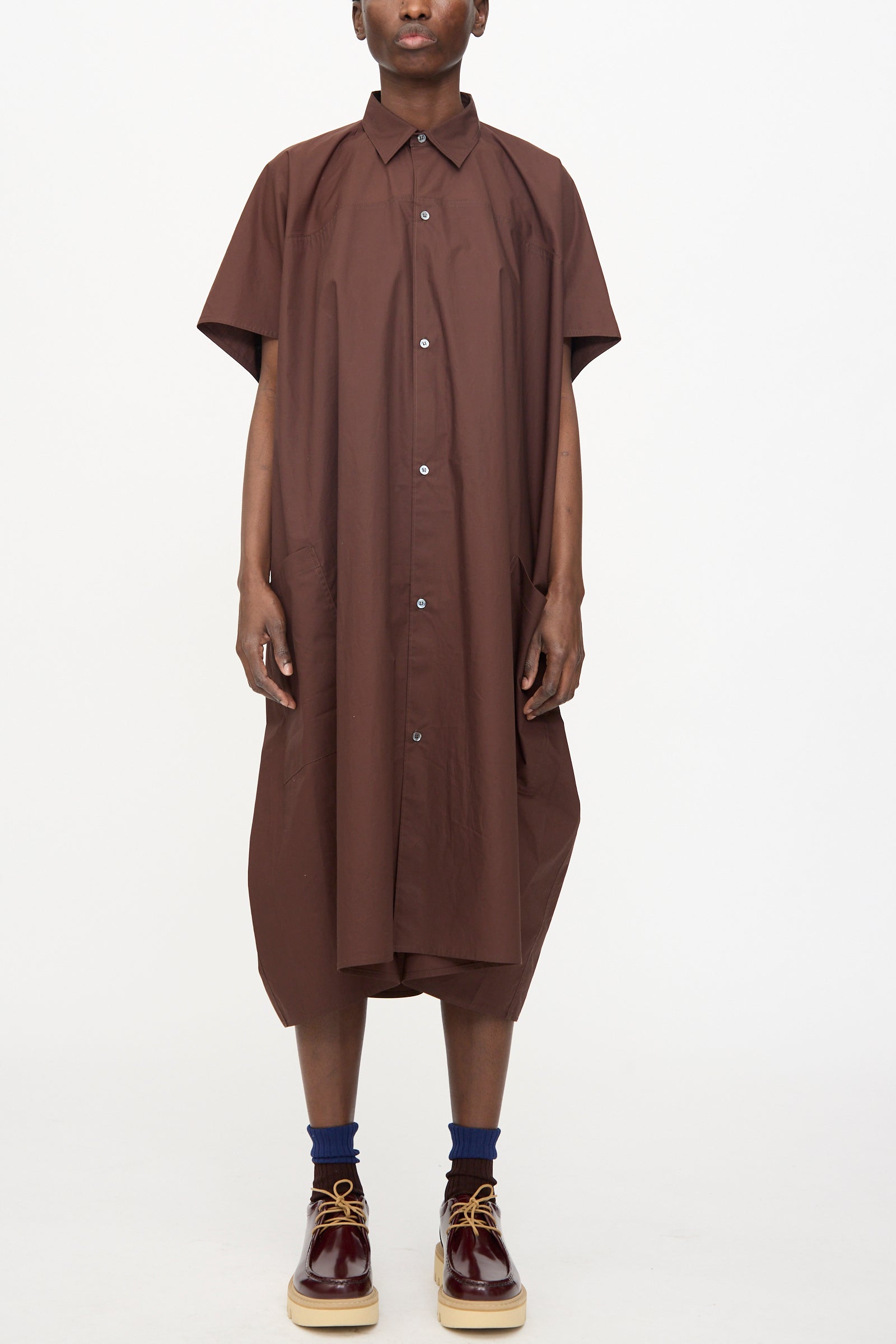 Model wears the Sofie D'Hoore Duchy Dress in Cacao—an oversized, short-sleeve brown cotton dress—with navy socks and burgundy lace-up shoes, standing against a plain white background.