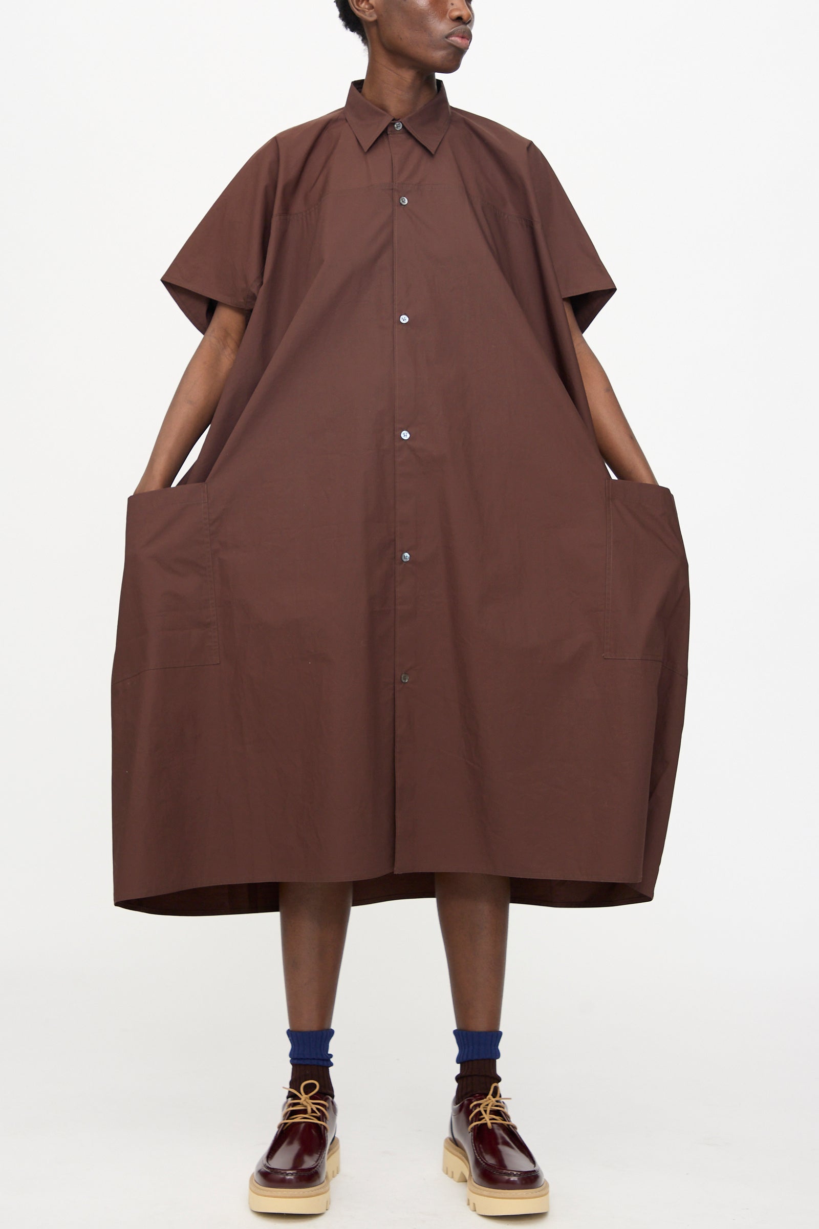 Model wears the Sofie D'Hoore Duchy Dress in Cacao, a loose-fit cotton midi dress with short sleeves and large front pockets, styled with dark socks and brown platform shoes against a plain white background.