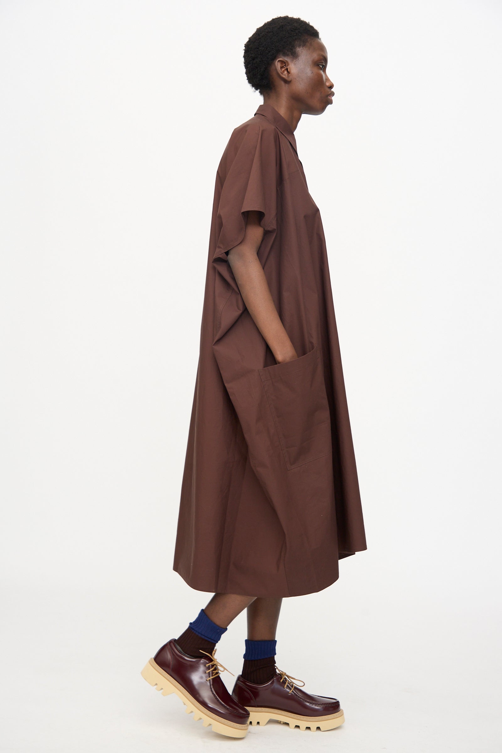 A person in profile wears the Sofie D'Hoore Duchy Dress in Cacao—an oversized cotton midi with short sleeves—styled with chunky maroon shoes and blue socks, set against a plain white background.