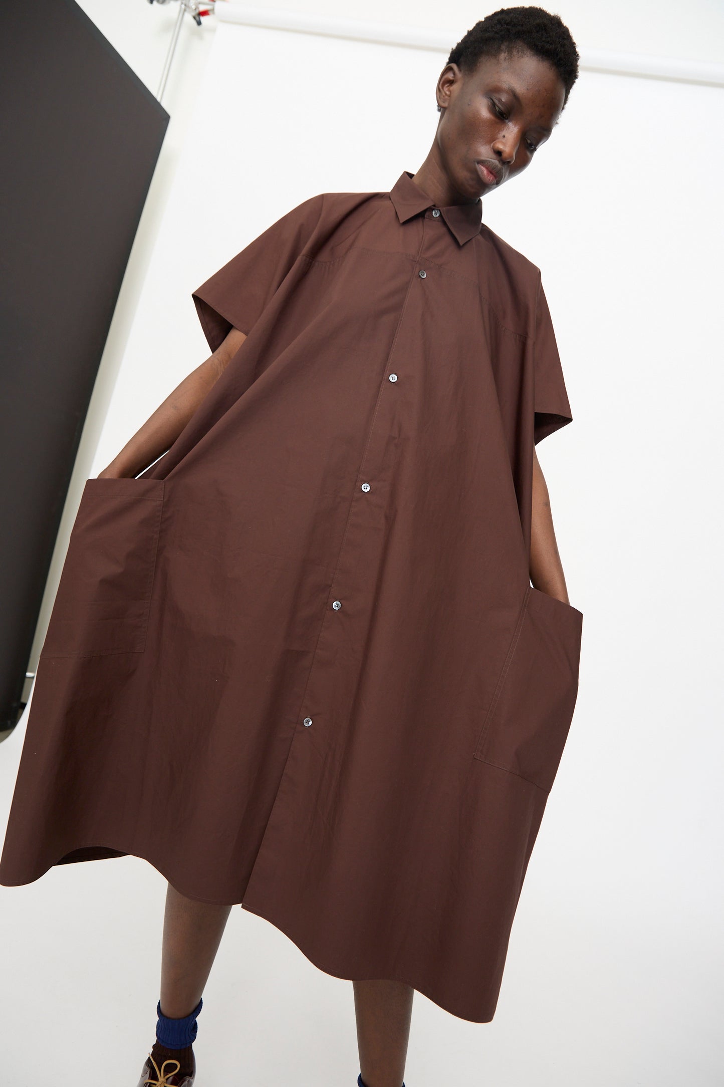 Model wears the Sofie D'Hoore Duchy Dress in Cacao, a loose, short-sleeved cotton dress with an oversized fit and pockets, standing against a plain studio background.