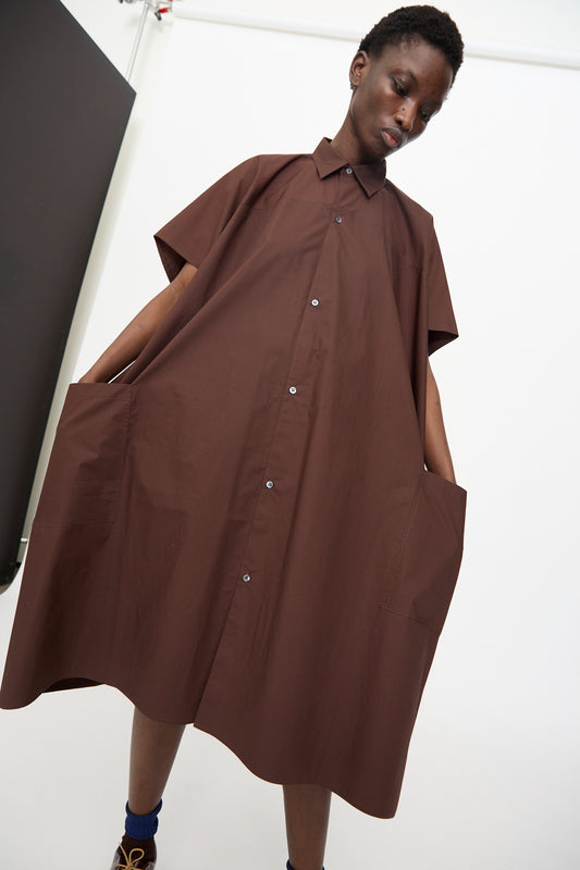 Model wears the Sofie D'Hoore Duchy Dress in Cacao, a loose, short-sleeved cotton dress with an oversized fit and pockets, standing against a plain studio background.