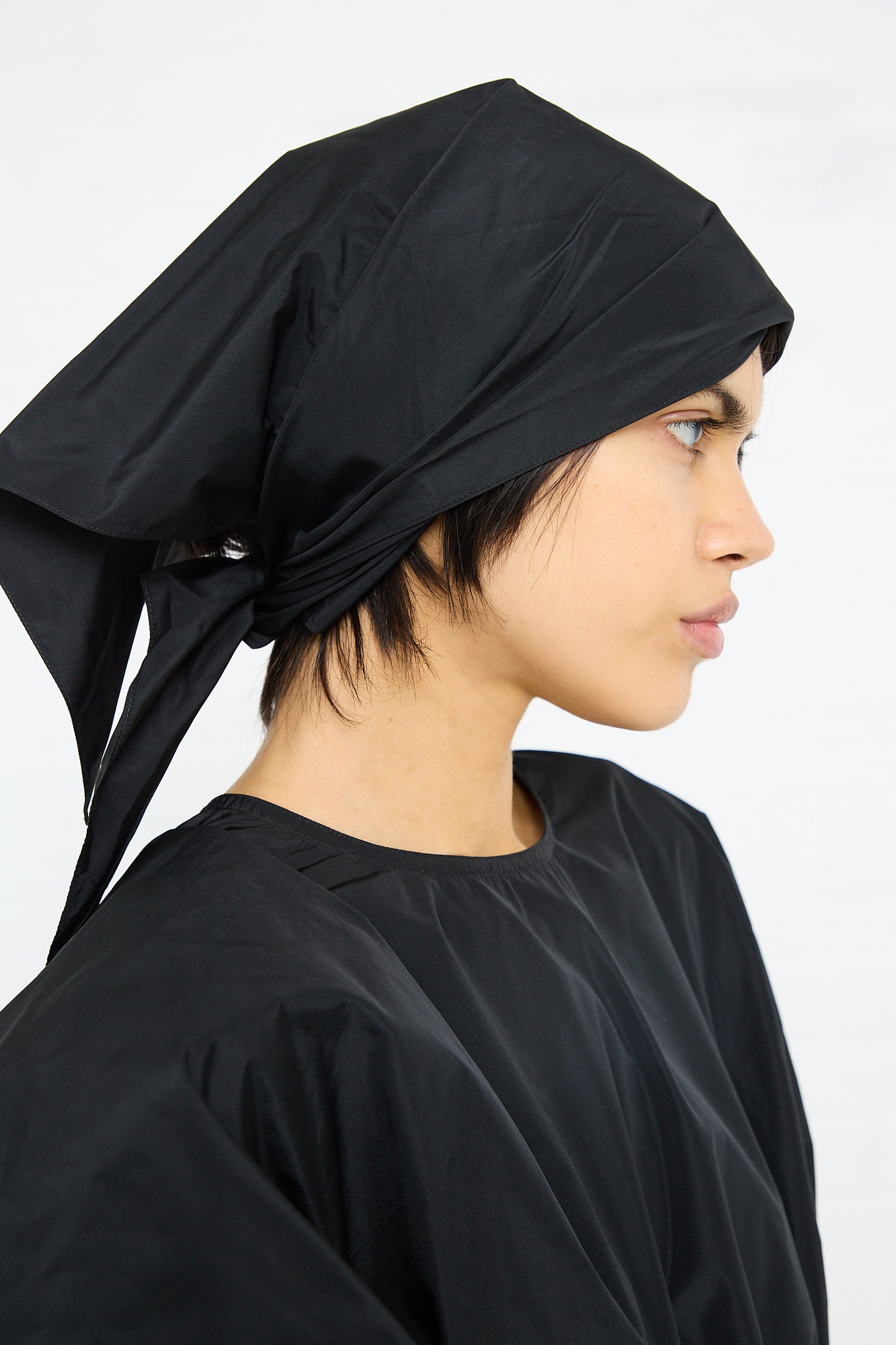 Profile view of a person wearing a Sofie D'Hoore Duke Dress with Lace Hem in Black headscarf and garment crafted from polyester taffeta against a white background.