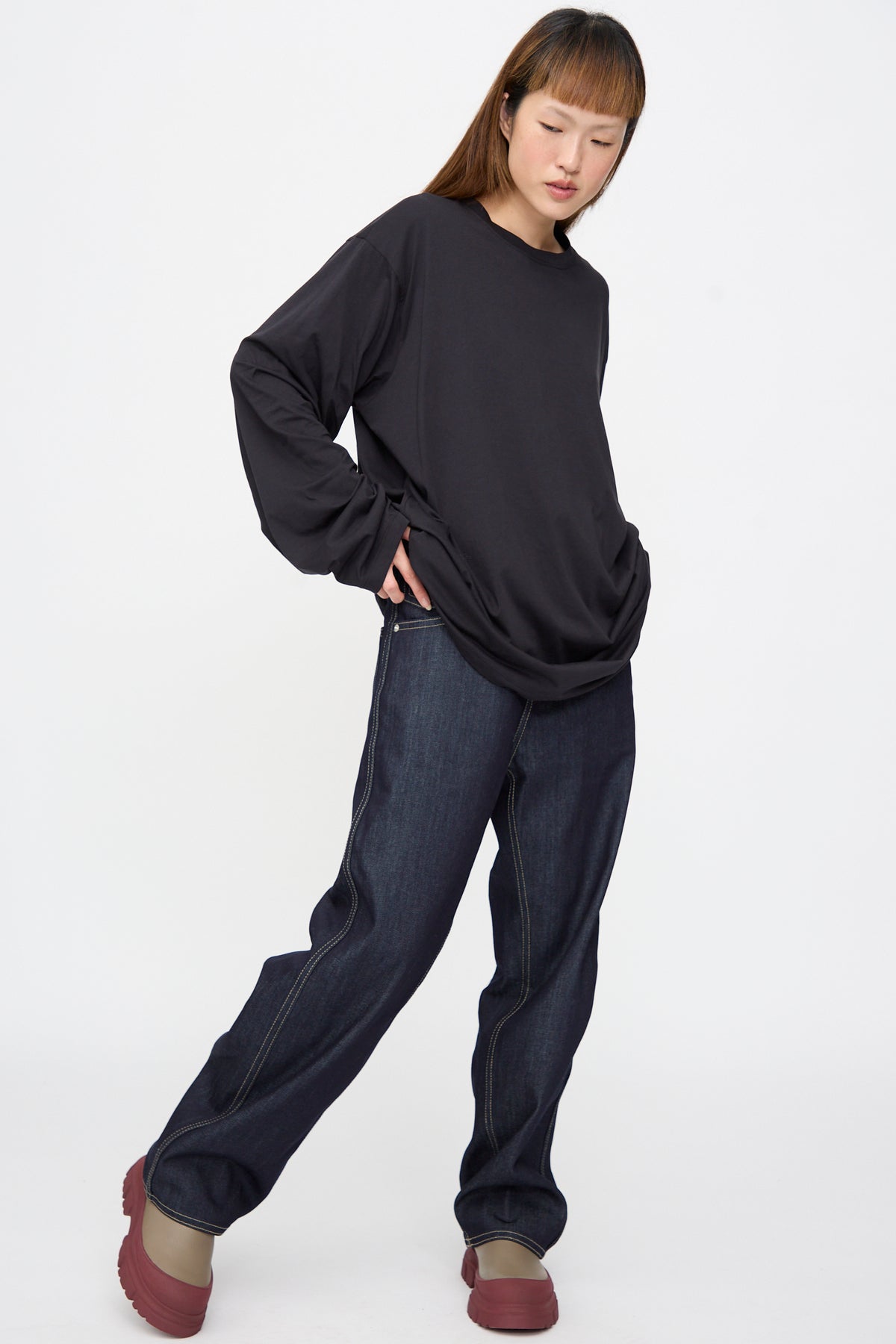 A person wearing an oversized, long-sleeve black Sofie D’Hoore Fine Cotton Jersey Toledo Tee in Cosmos, dark denim jeans, and brown shoes with thick, red platform soles.