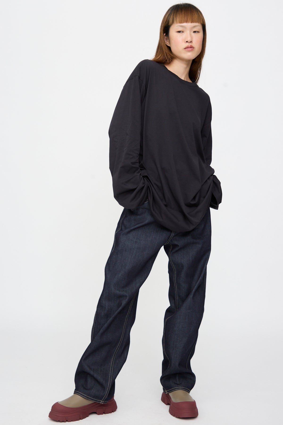 A person wearing a black Sofie D’Hoore Fine Cotton Jersey Toledo Tee in Cosmos, dark denim jeans, and olive-colored shoes with thick red lug soles.