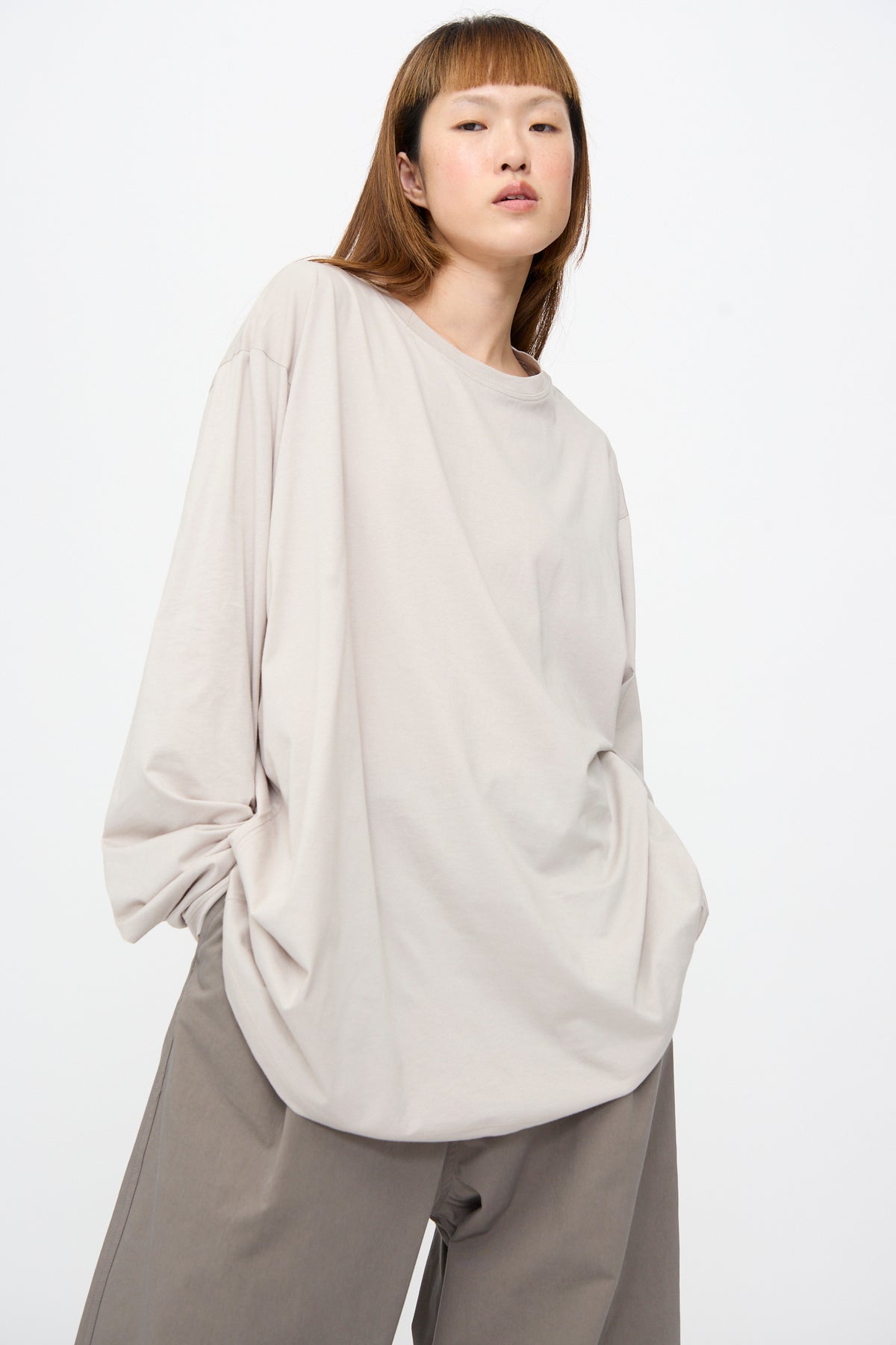 A model wearing a Sofie D’Hoore Fine Cotton Jersey Toledo Tee in Powder, an oversized, light beige long-sleeve cotton jersey crew neck t-shirt, and grey trousers.