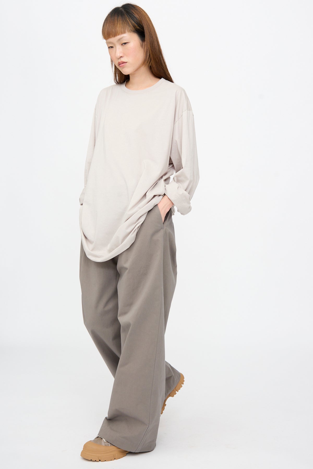 A person wearing an oversized Fine Cotton Jersey Toledo Tee in Powder and grey wide-leg trousers, with one hand in their pocket.