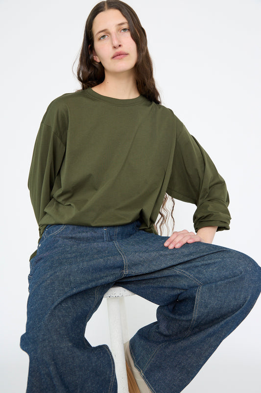 A person with long hair sits on a stool, modeling the Sofie D'Hoore Fine Cotton Jersey Toledo Tee in Seaweed and wide-leg jeans against a plain background.