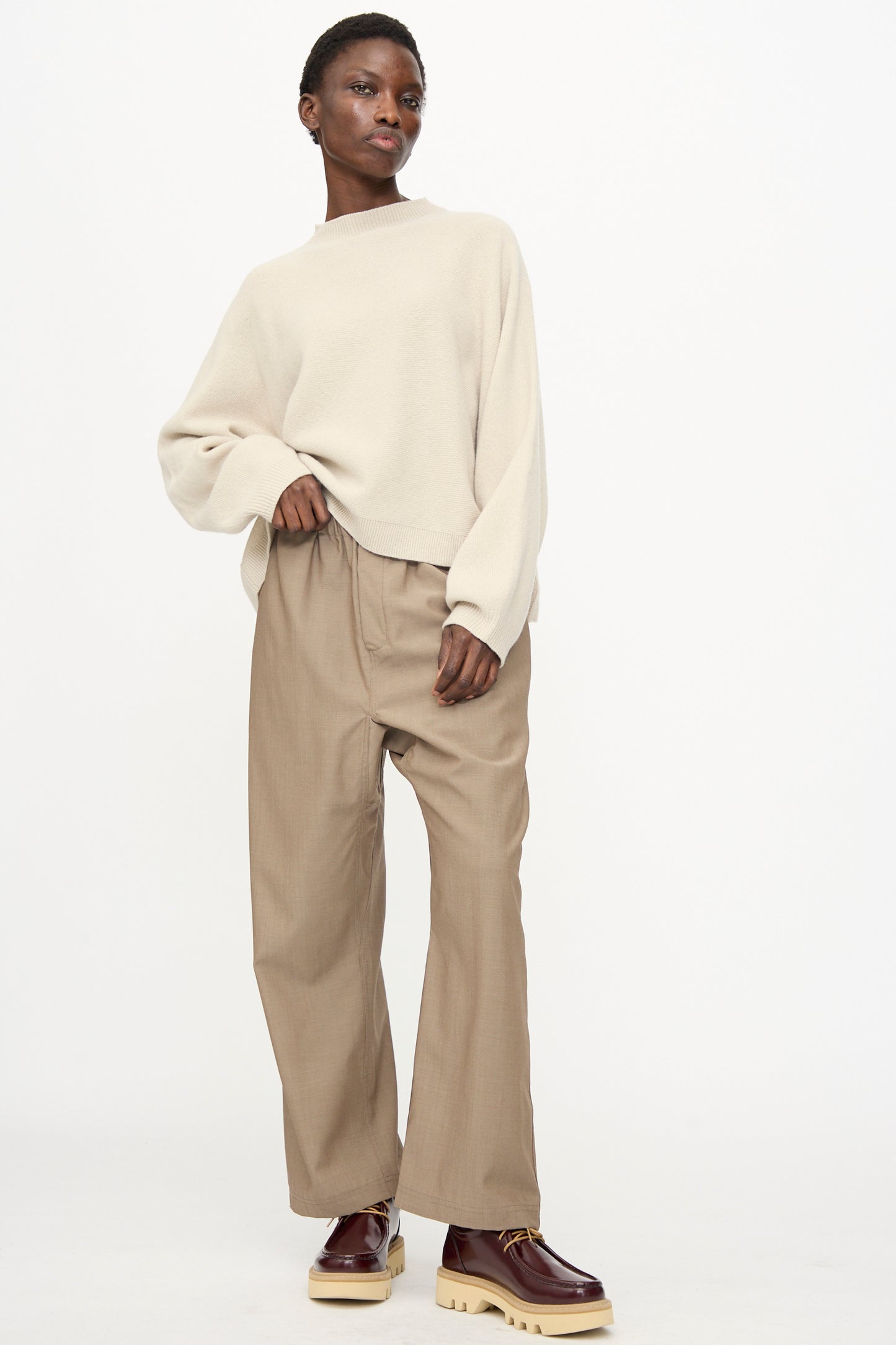 A person stands against a plain background wearing a loose beige sweater, Sofie D'Hoore's Light Mohair Wool Pinot Pant in Quartz, and brown shoes with chunky soles.
