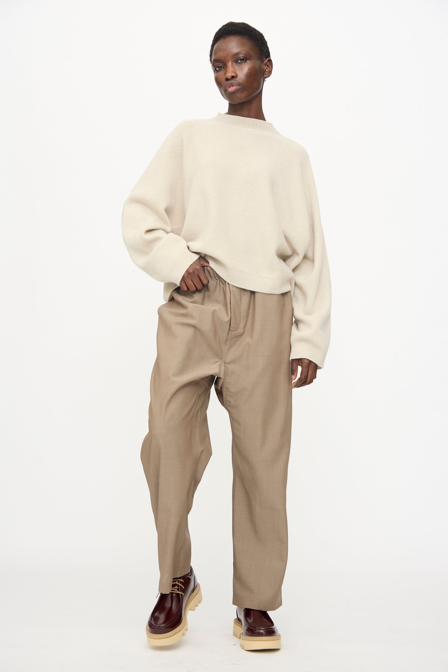 A person stands against a white background, wearing a loose beige sweater, Sofie D'Hoore's Light Mohair Wool Pinot Pant in Quartz, and burgundy platform shoes.