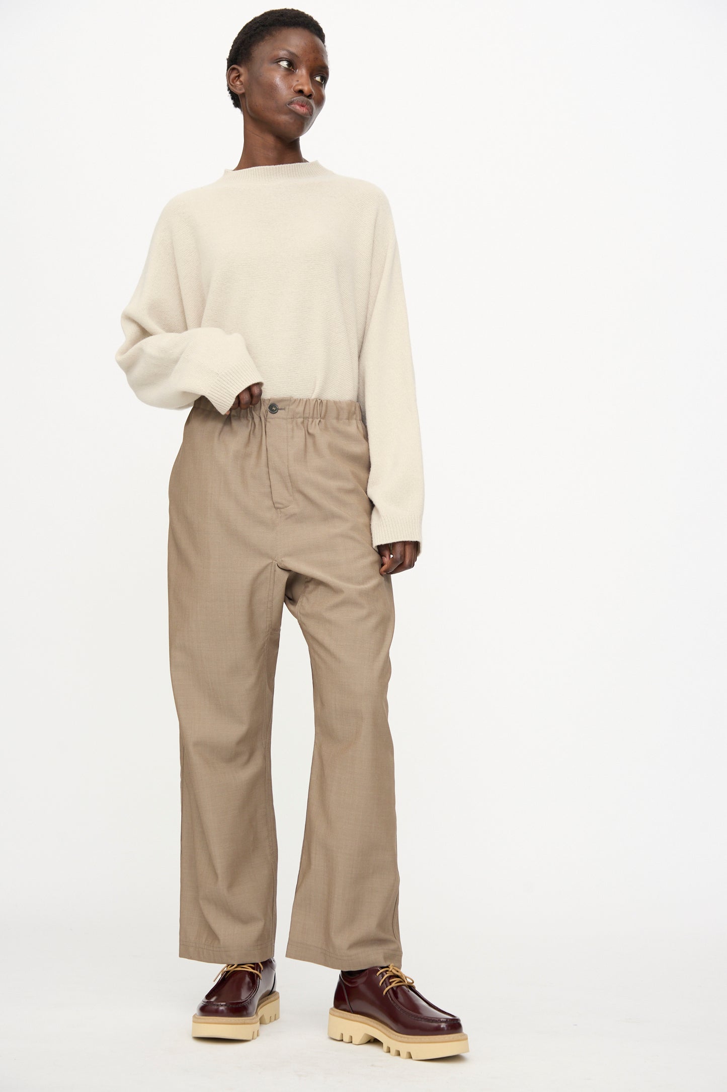 A person stands against a plain background wearing a cream sweater and Sofie D'Hoore's Light Mohair Wool Pinot Pant in Quartz, paired with brown platform shoes.