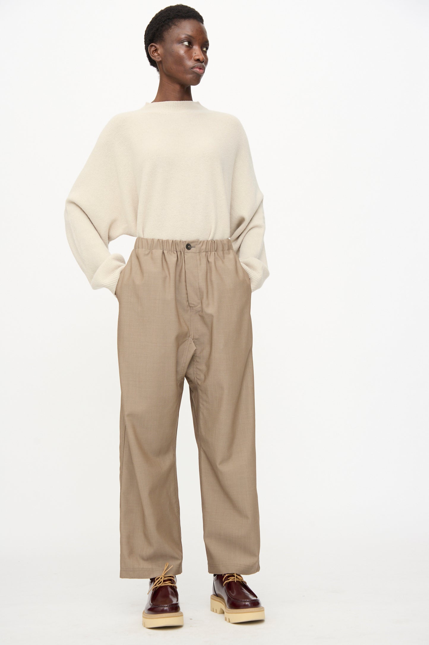 A person stands against a plain background wearing relaxed fit Sofie D'Hoore Light Mohair Wool Pinot Pants in Quartz, a loose beige sweater, and brown platform shoes.