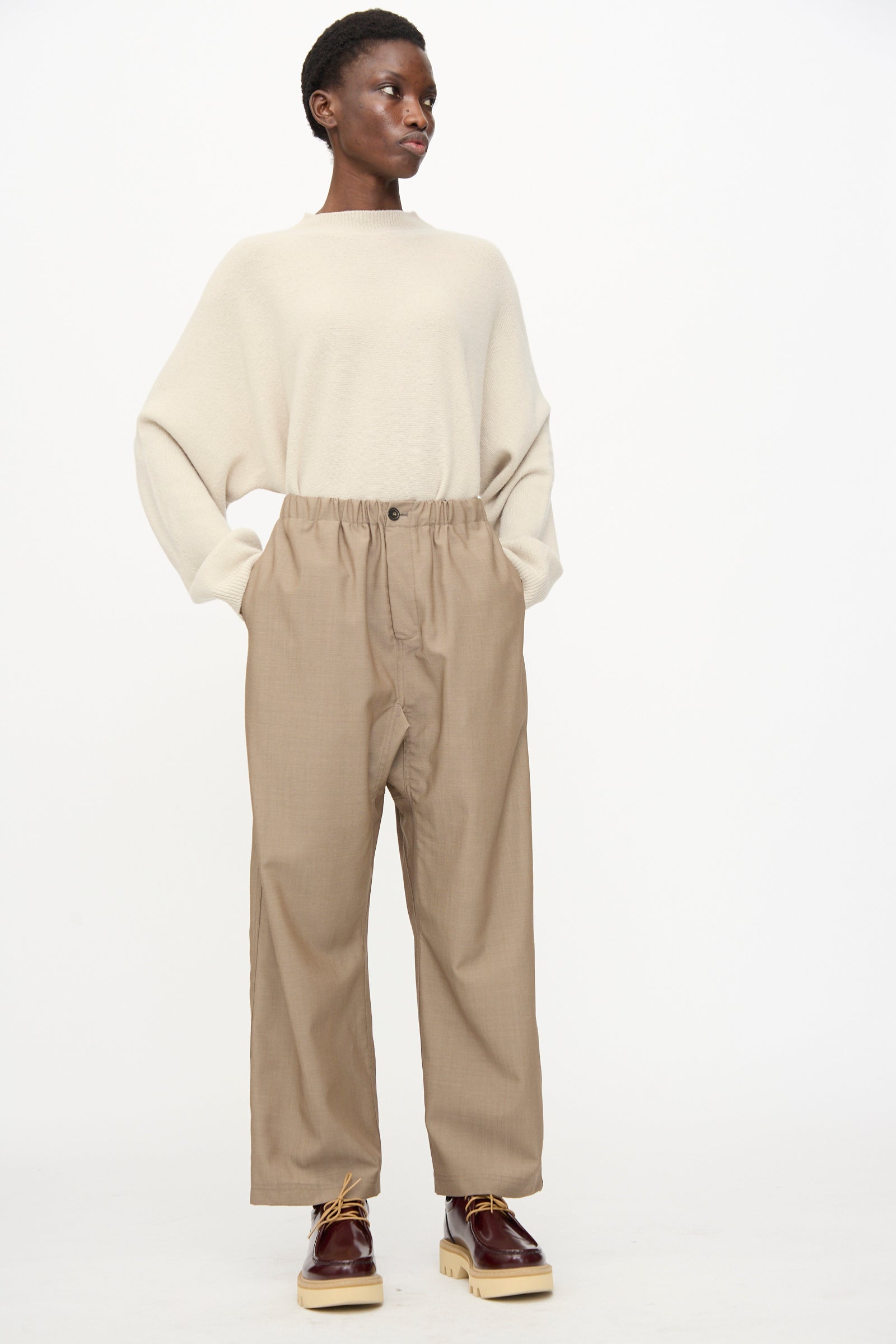 A person stands against a plain background wearing relaxed fit Sofie D'Hoore Light Mohair Wool Pinot Pants in Quartz, a loose beige sweater, and brown platform shoes.