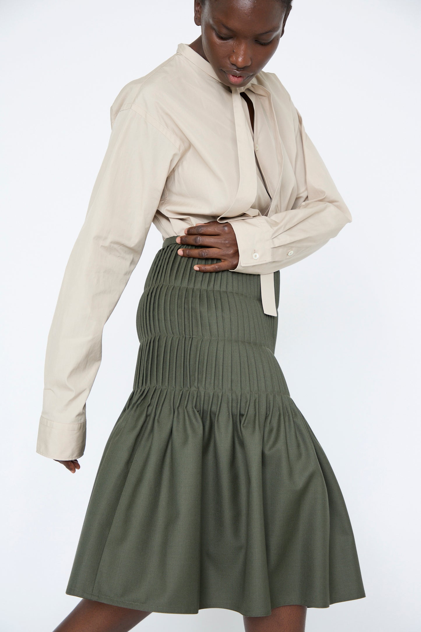 A woman wearing a beige button-up shirt and the Light Worsted Wool Shake Pleated Skirt in Fern by Sofie D'Hoore, posing against a plain background, walking to the left.
