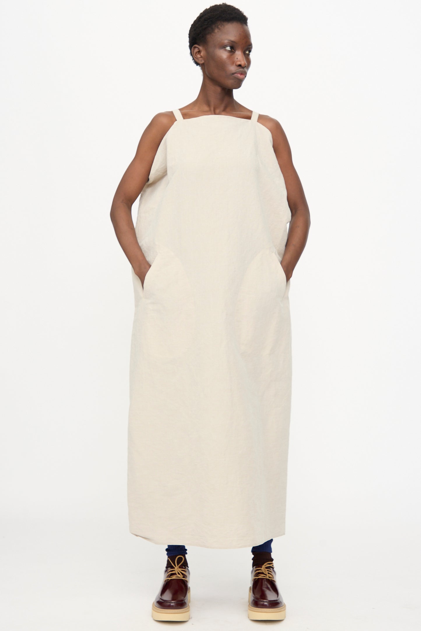A person stands against a plain background wearing the Sofie D'Hoore Linen Cotton Dele Dress in Natural, a loose, sleeveless maxi dress with large pockets, paired with dark pants and brown lace-up shoes.