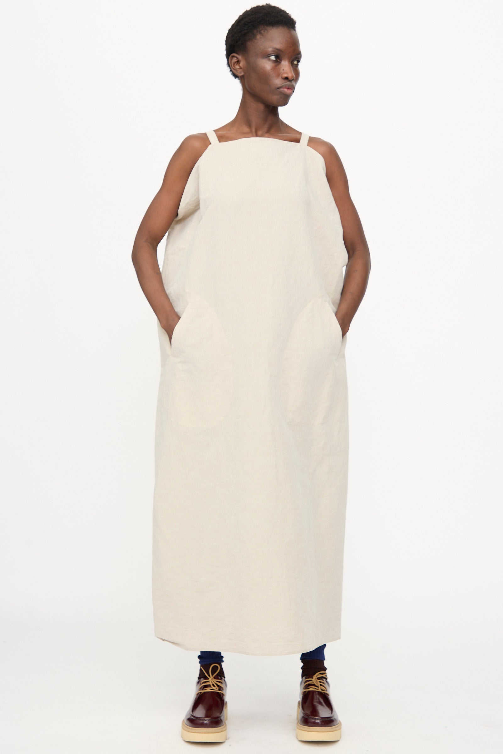 A person stands against a plain background wearing the Sofie D'Hoore Linen Cotton Dele Dress in Natural, a loose, sleeveless maxi dress with large pockets, paired with dark pants and brown lace-up shoes.