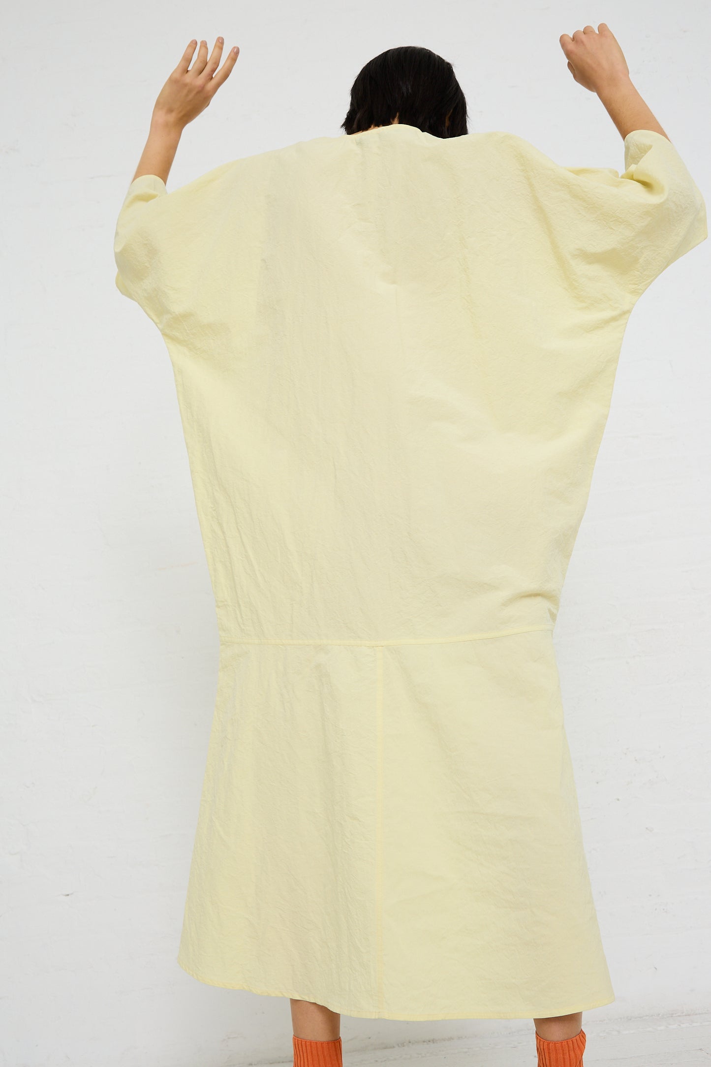 Person standing with their back to the camera, raising their arms, wearing a pale yellow oversized Linen Cotton Delfi Dress in Pastis by Sofie D'Hoore.