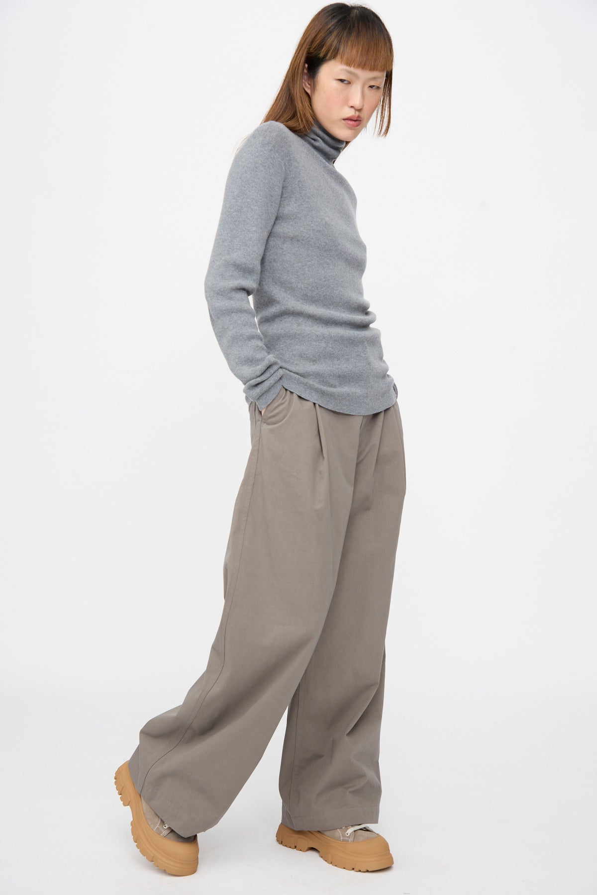 A person wearing a grey Malika Sweater in Grey Melange rib-knit turtleneck, wide-leg trousers, and tan lug-sole shoes.