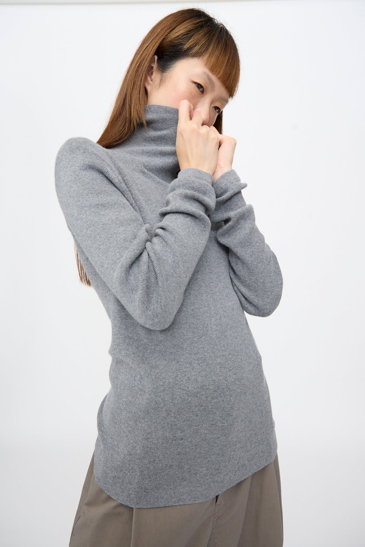 A person wearing a grey rib-knit Malika Sweater in Grey Melange, holding the turtleneck near their face.