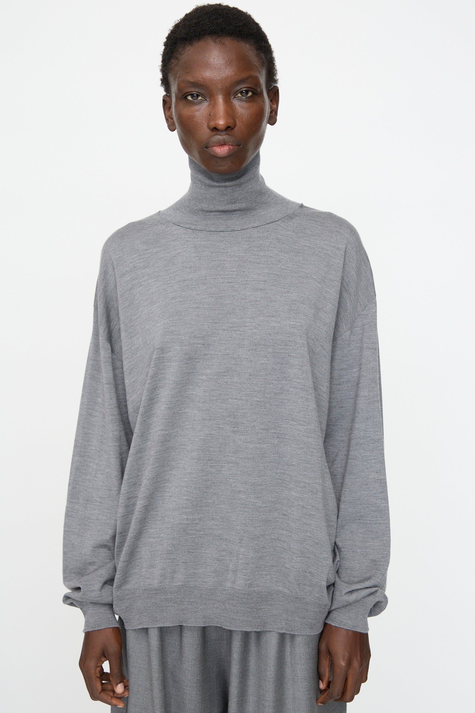 A person wearing the Sofie D'Hoore Merino Maffei Sweater in Steel Melange stands against a plain white background, paired with matching pants.