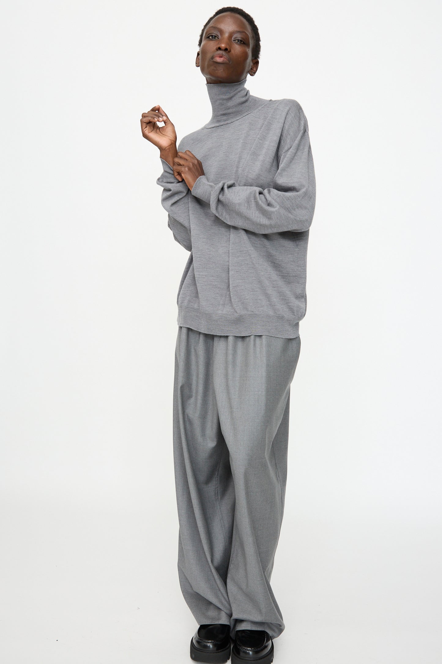 A person wears the Sofie D'Hoore Merino Maffei Sweater in Steel Melange and wide-leg grey trousers, standing against a white background. 