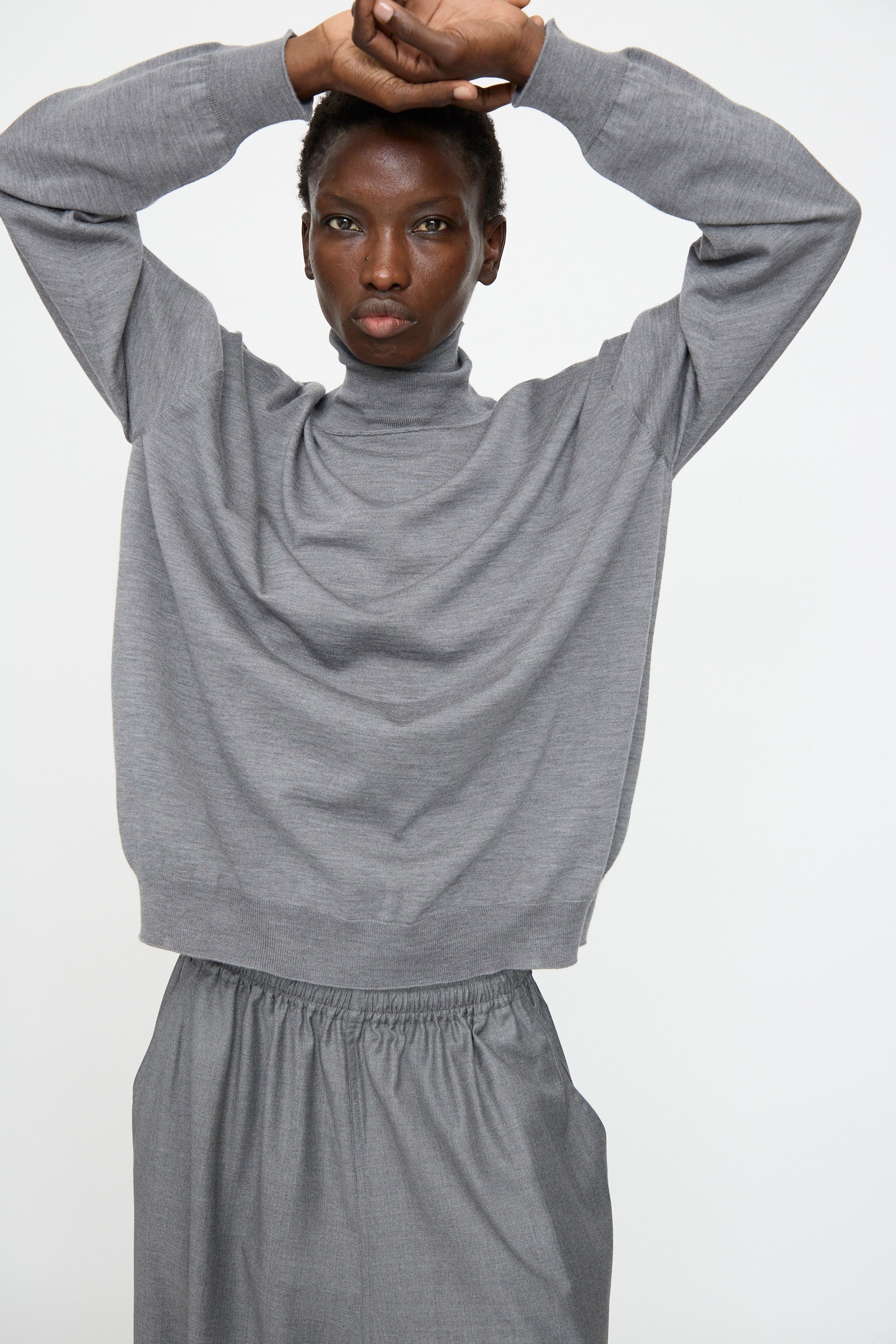 A person wears the Sofie D'Hoore Merino Maffei Sweater in Steel Melange with a matching grey skirt, standing with arms crossed above their head against a plain background.