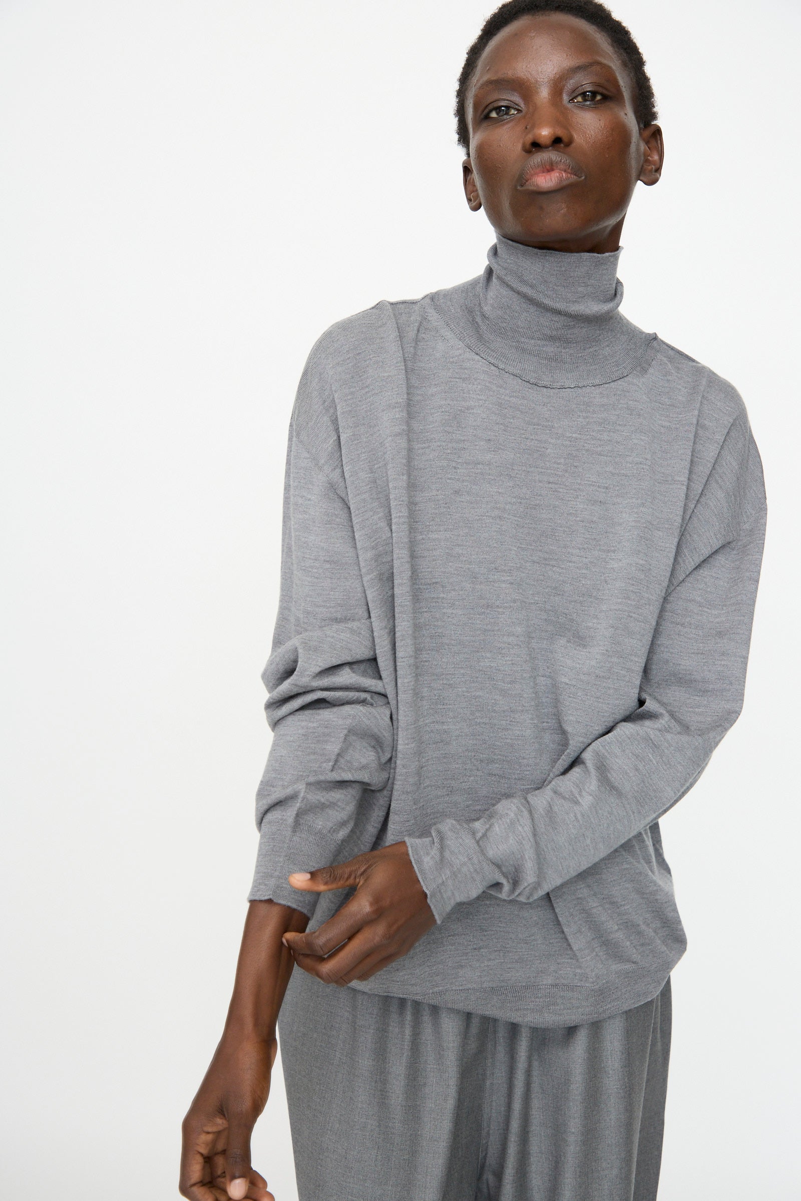 A person wears the Sofie D'Hoore Merino Maffei Sweater in Steel Melange with matching gray pants, standing against a plain light background and slightly adjusting their sleeve.