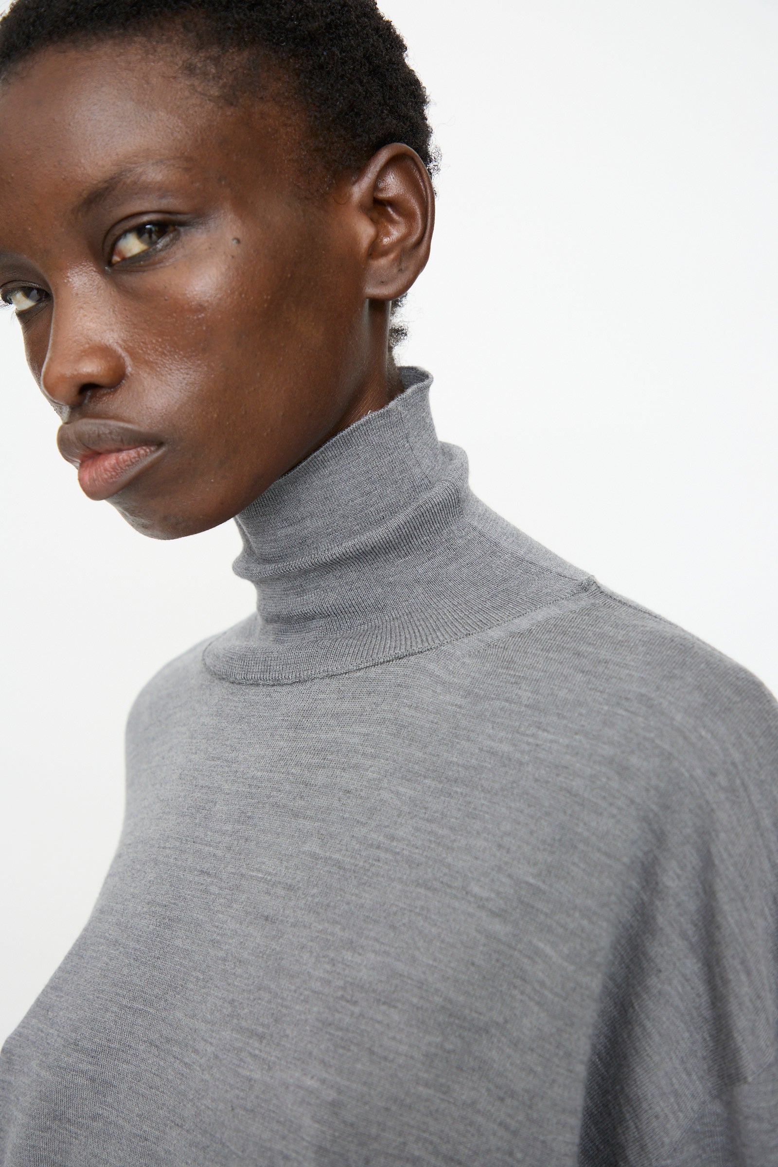 A person wears the Sofie D'Hoore Merino Maffei Sweater in Steel Melange, looking slightly to the side against a plain white background.