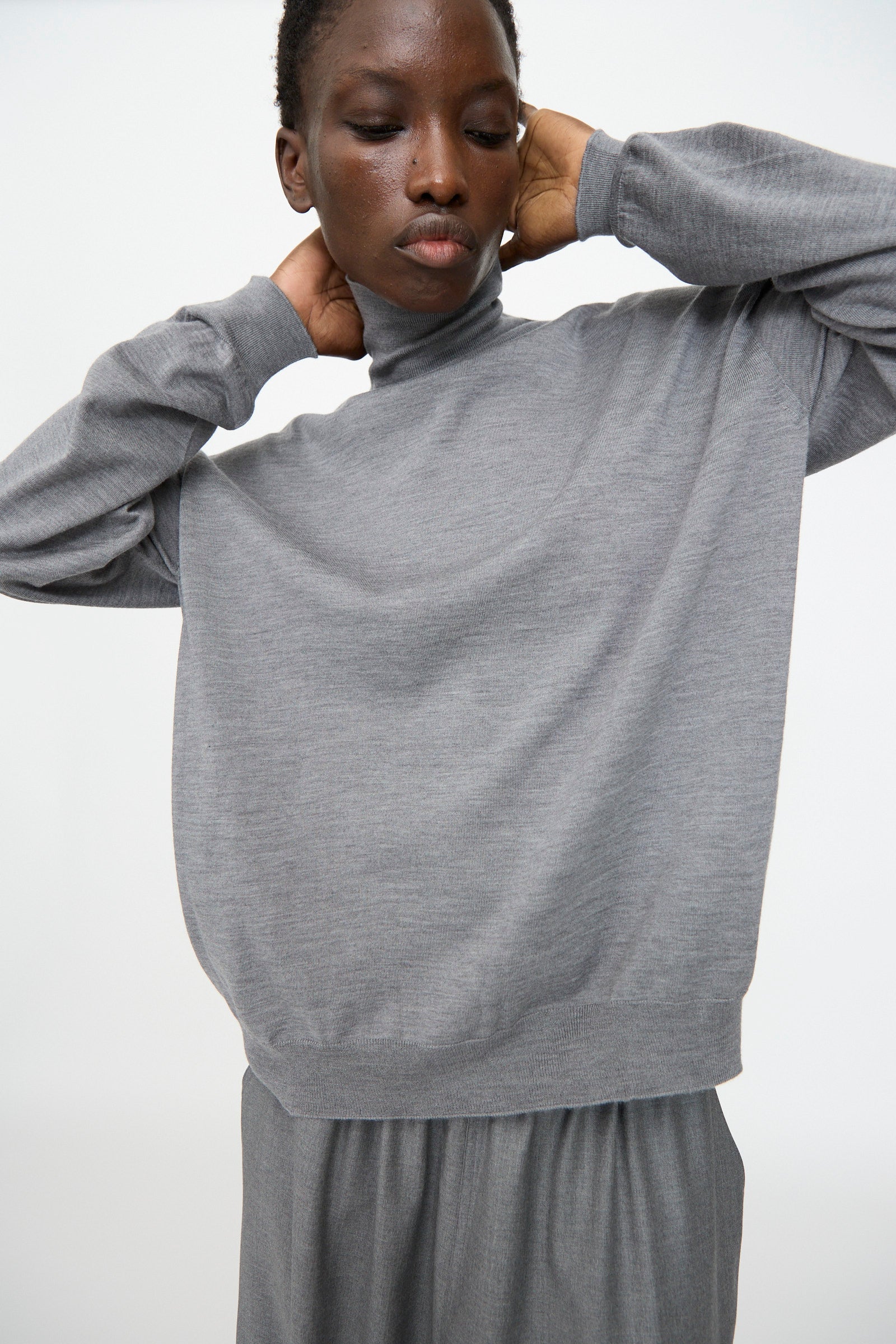 A person wears the Sofie D'Hoore Merino Maffei Sweater in Steel Melange, a loose-fitting extra fine merino wool turtleneck, paired with matching pants and posing against a plain light background with hands touching their neck.