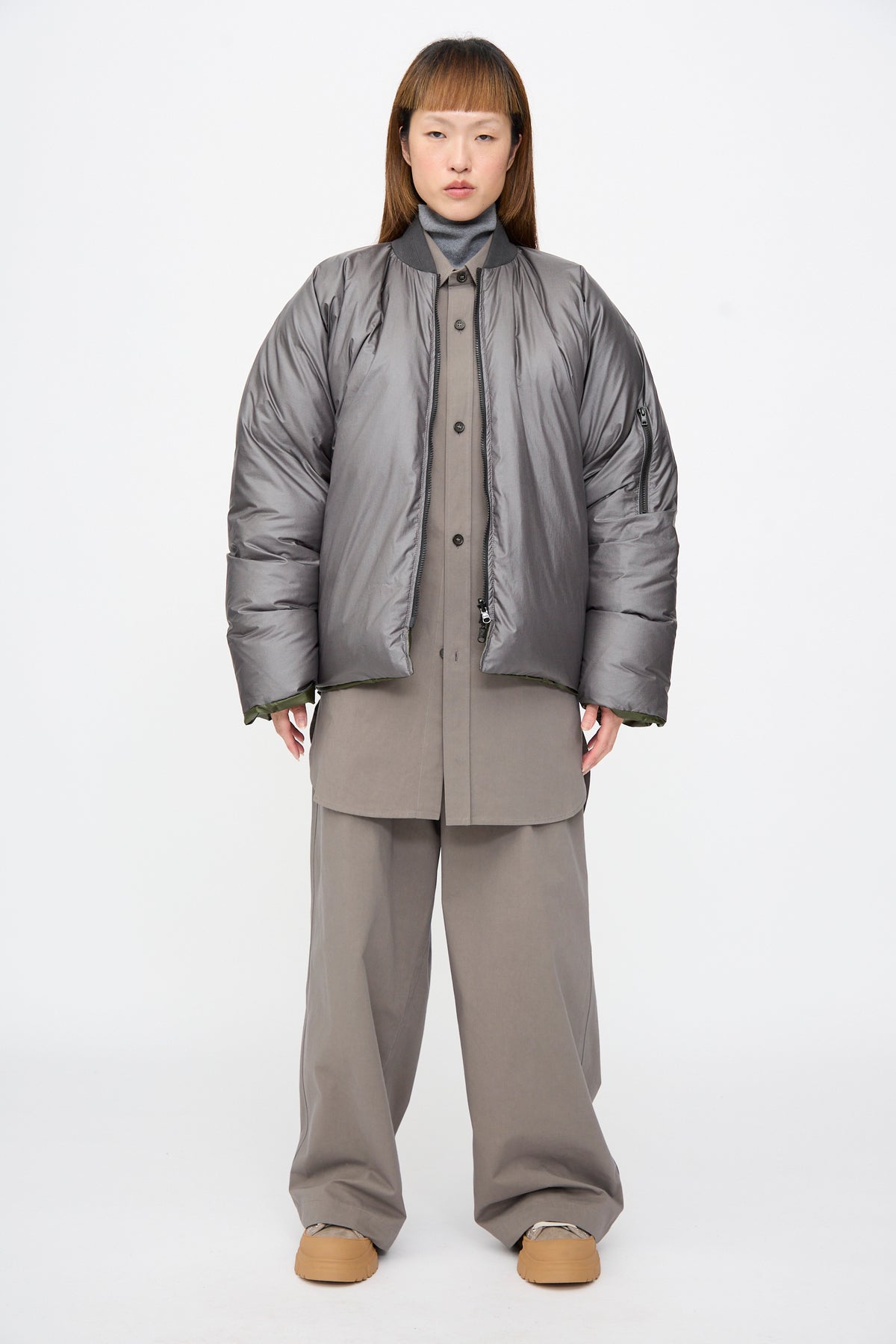 A person wearing the Sofie D’Hoore Origin Down Jacket in Charcoal and Khaki over a khaki button-down shirt and matching wide-leg trousers.
