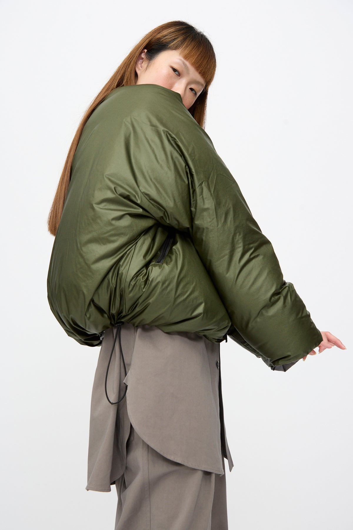 A person wearing an olive green, oversized fit Sofie D’Hoore Origin Down Jacket in Charcoal and Khaki with a cinched waist and grey trousers.