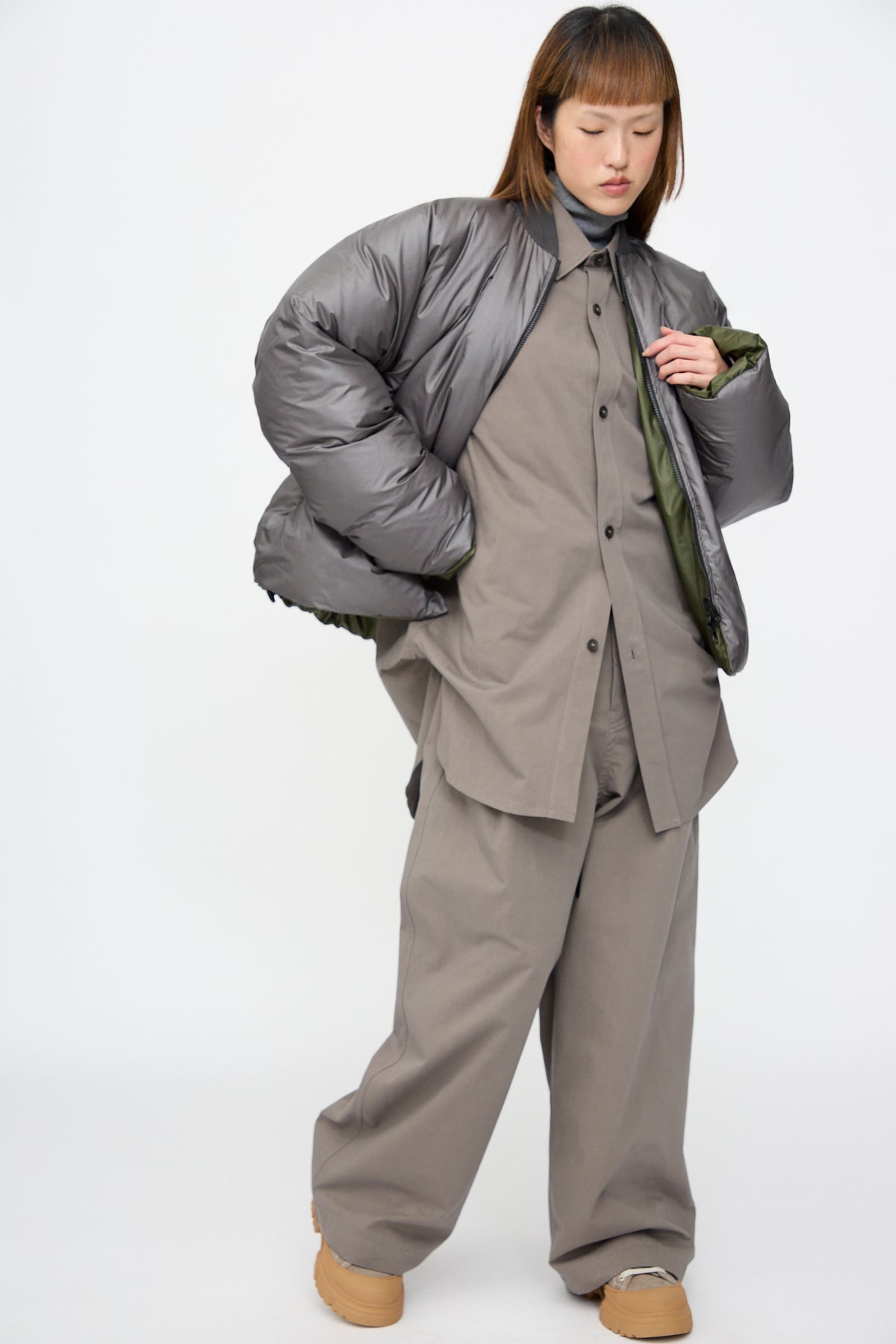 A person wearing a reversible Sofie D’Hoore Origin Down Jacket in charcoal and khaki over a matching grey button-up shirt and trousers with tan platform shoes.