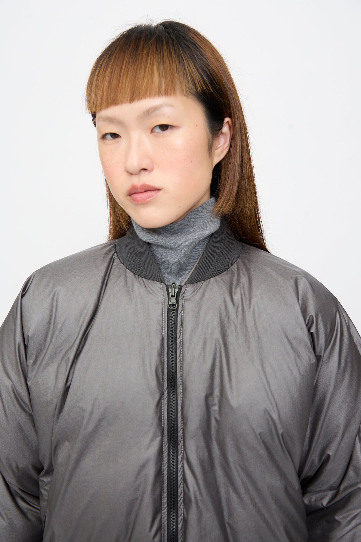 A person wearing an oversized, charcoal gray and khaki Sofie D’Hoore Origin Down Jacket, a reversible zip-up puffer bomber, over a gray turtleneck sweater.